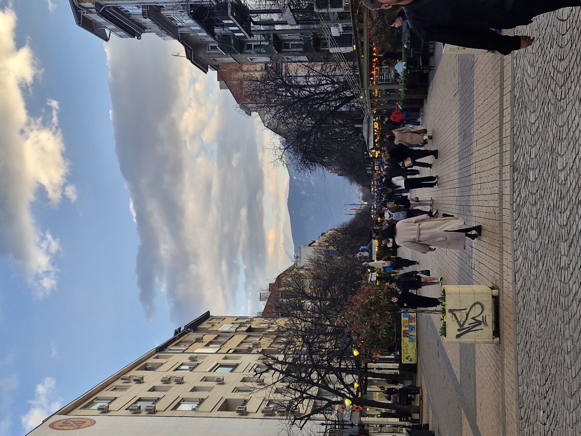 The image depicts Vitosha Boulevard in Sofia, Bulgaria, looking southeast towards the snow-capped Vitosha Mountain. The scene is a moderately crowded pedestrian street lined on both sides by multi-story buildings under a partly cloudy sky, with light suggesting late afternoon or early evening.
On the left, a light-colored, multi-story building features numerous external air conditioning units and balconies. A large, circular red and yellow logo, resembling the number "16", is prominently displayed near the top left corner of this building. Further down the left side, several bare deciduous trees with dark branches line the boulevard, interspersed with street lamps emitting a warm, yellow light. Ground-level establishments along this side have outdoor seating areas.
On the right, buildings display older architecture, some with exposed brick and multiple balconies, continuing down the street. These buildings also host ground-level businesses with illuminated outdoor dining setups, featuring tables, chairs, and overhead awnings or string lights.
The pedestrian walkway in the foreground is paved with dark cobblestones, transitioning to a pattern of light and dark rectangular pavers further into the mid-ground. A light-colored, rectangular concrete planter with graffiti, specifically stylized "NKS" in dark spray paint, containing yellow flowers, is situated on the left side of the walkway.
Numerous individuals are traversing the boulevard. In the immediate foreground, the right side of a person wearing a dark top is visible, walking towards the viewer. In the mid-ground, a female wearing a light beige long coat walks away from the viewer, centered on the main path. Other people, dressed in seasonal outerwear like jackets and coats, walk in various directions, individually or in small groups. Some are seated at the outdoor dining areas. In the distant background, the silhouette of snow-covered mountains, identifiable as Vitosha Mountain, is visible, capped by clouds.