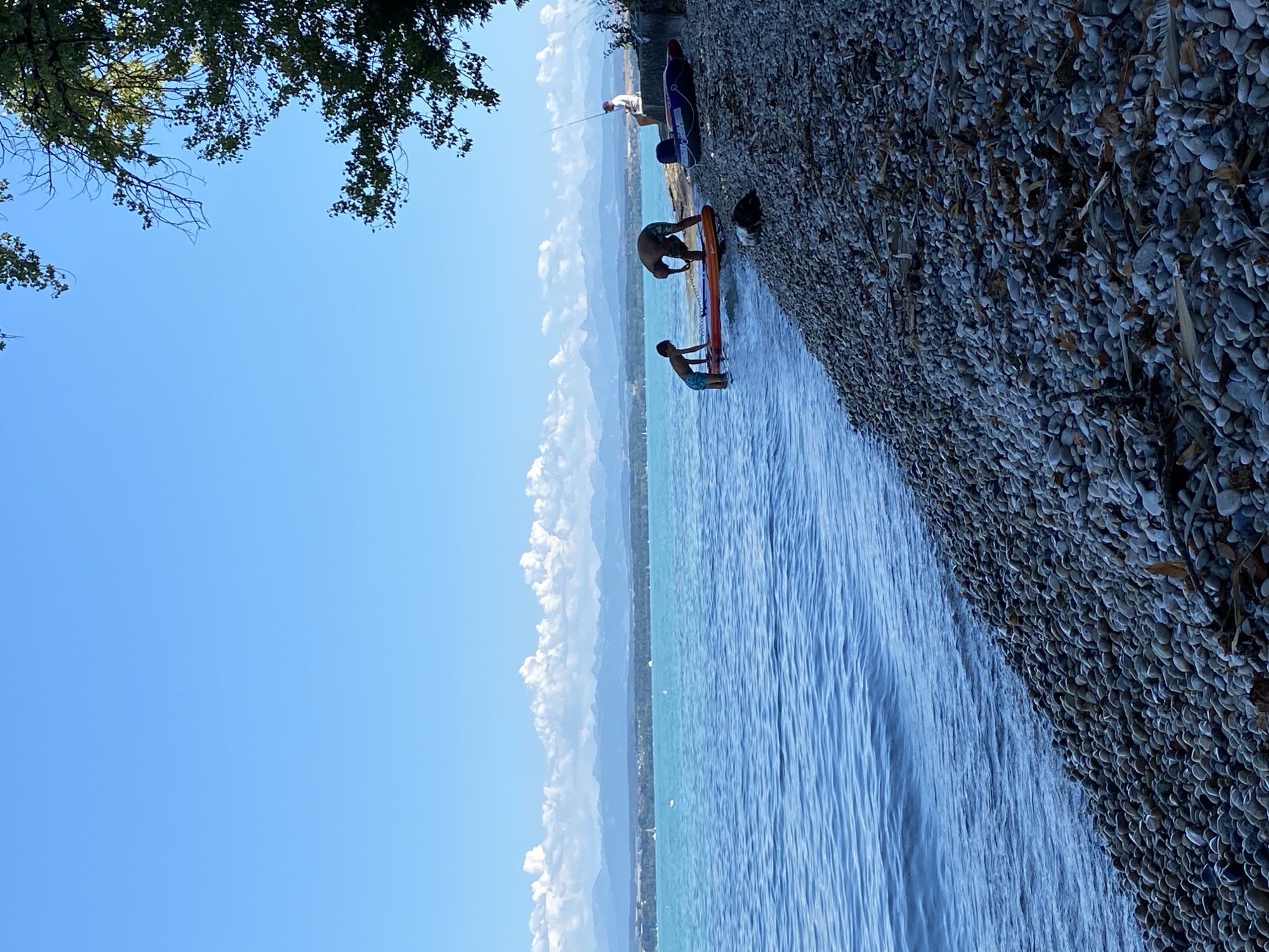 In Cortaillod, Switzerland, on a sunny day, a pebble beach meets a bright blue lake. In the foreground, the shore is covered with smooth, grey and white pebbles, interspersed with some dry leaves and twigs. The lake's surface is calm with gentle ripples, reflecting the clear sky.
Further out in the lake, two people are interacting near the water's edge. One individual, a child wearing blue shorts, stands in the shallow water, appearing to hold onto an orange paddleboard. Beside them, an adult, wearing camouflage shorts, bends over, possibly adjusting or preparing the paddleboard.
On the shore to the right, a person is seated on a concrete wall, holding a fishing rod and fishing in the lake. Nearby, on the same wall, are two inflatables: a dark blue and red one, and a smaller, darker one. Behind them, the edge of a large tree with green leaves frames the upper right corner of the image.
In the distance, across the lake, a long, low-lying landscape is visible, featuring trees and buildings, backed by a range of mountains. The sky is a vibrant, cloudless blue, with a few wispy clouds appearing near the horizon above the distant mountains.