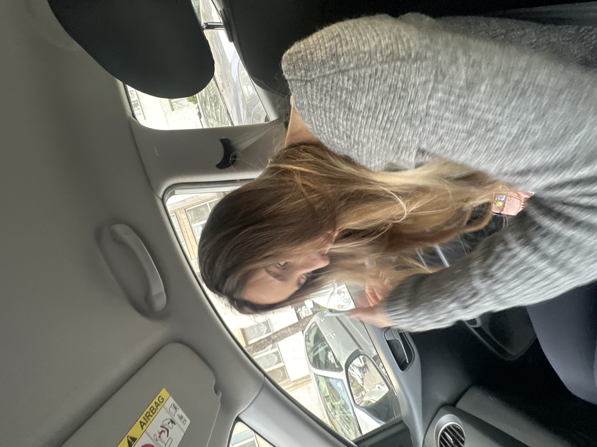 A person is inside a car, possibly in Sofia, Bulgaria, though this is not directly verifiable from the image. The individual has long brown hair and is wearing a grey sweater. They are looking downwards and towards the side of the car, possibly interacting with something outside the frame. The interior of the car is visible, including the airbag warning on the sun visor, a passenger side grab handle, the car's window, a headrest, and a dashboard with an air vent. Outside the car window, there are buildings and other vehicles.