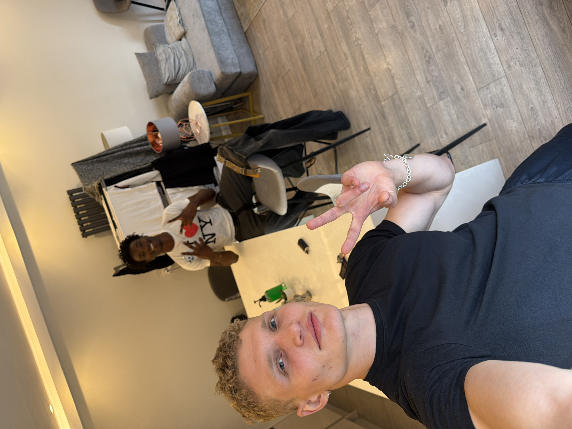 The image depicts two young men indoors, likely in a modern apartment. The man in the foreground, with blonde curly hair, is wearing a black t-shirt and making a peace sign with his right hand. He has a silver hoop earring in his left ear and a silver bracelet on his right wrist. He is positioned in front of a white table. Behind him, another man with dark curly hair, wearing a white t-shirt with "I ❤️ NY" printed on it and dark pants, is seated on a stool. He is also making a peace sign with both hands. The room features a grey sofa with pillows, a round side table with a lamp, and a radiator. The flooring is light-colored wood. The overall setting suggests a casual, domestic environment. The image does not contain any specific indicators of its location within the city of London, United Kingdom.