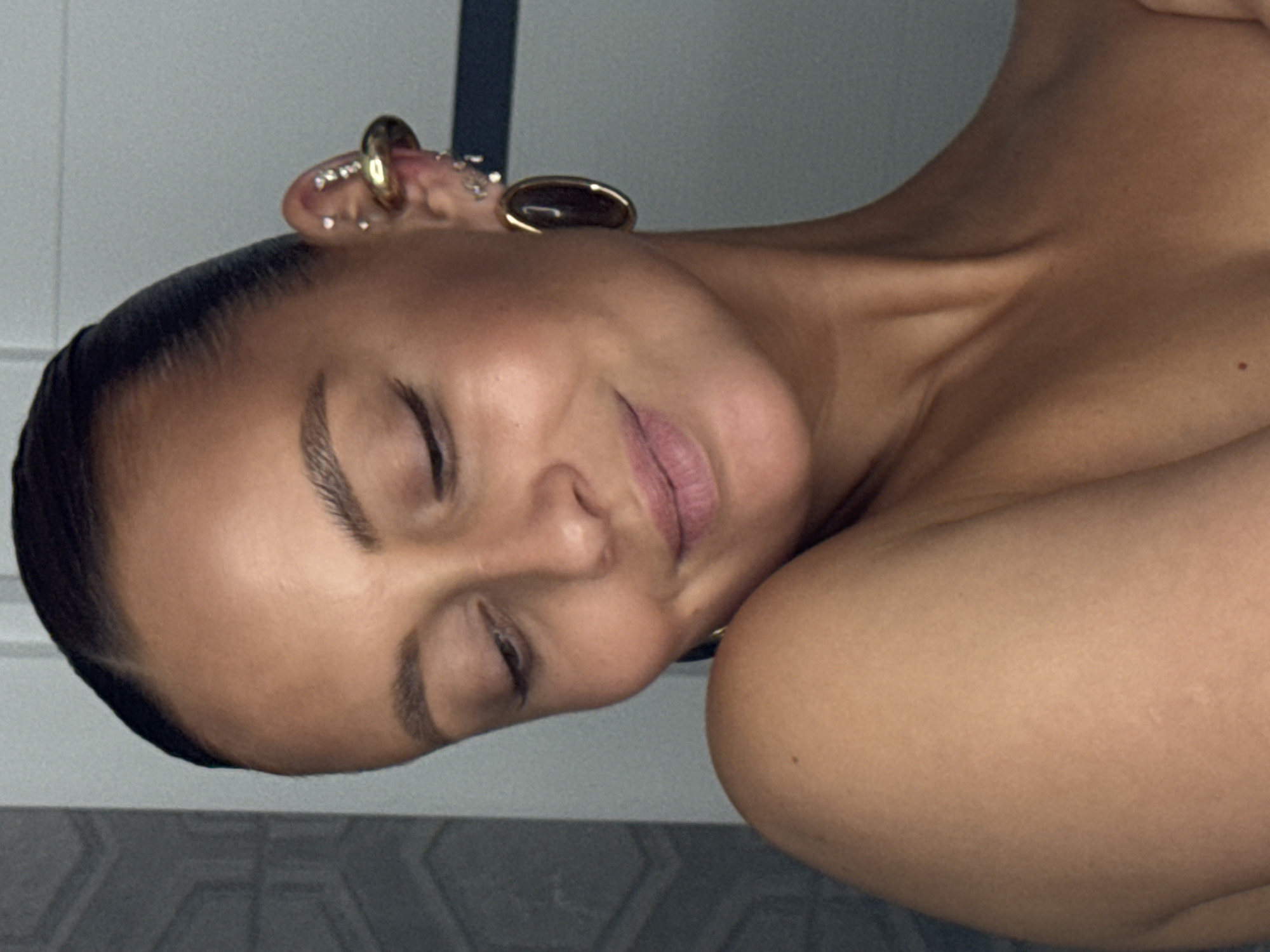 A close-up portrait depicts a person, likely female, from the upper chest upwards, with tanned skin and dark hair tightly pulled back, revealing a clean hairline. The individual's head is tilted downwards, eyes are closed or heavily downcast, and a subtle, closed-mouth smile is visible. The right ear (viewer's left) features multiple piercings, including small clear studs along the helix, a prominent gold-toned ear cuff, and a large oval hoop earring with a dark brown/black center in the lobe. Bare shoulders and neck are visible, with a small dark mole on the right shoulder (viewer's left). The background consists of a light grey wall with a faint geometric pattern on the left and a plain light grey surface on the right, bisected by a dark vertical line, indicating an indoor environment. The scene is located in the city of Chaikovsky, Russia.