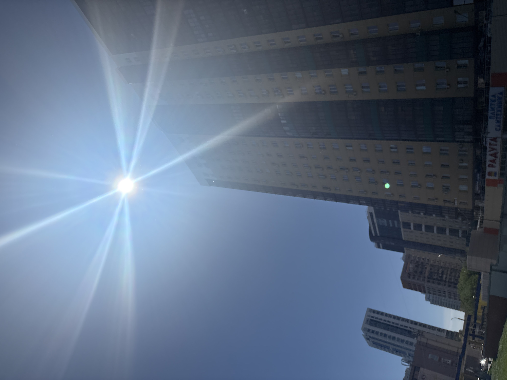 The image shows a view looking up at several tall buildings in Perm, Russia, with the sun in the upper left casting bright rays across the sky. The tallest building is a modern skyscraper with many windows and dark, reflective glass, potentially an office building or residential tower. Other buildings are visible in the midground and background, varying in height and architectural style. A sign with Russian text is partially visible attached to the side of the largest building. The sky is clear blue.