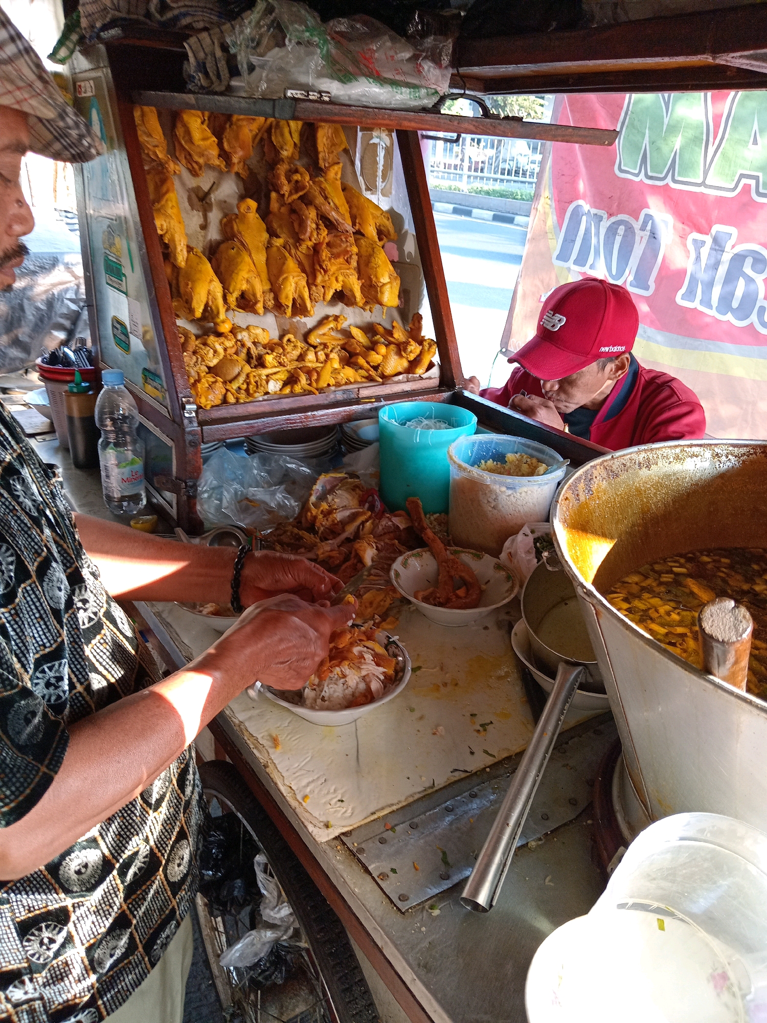 In Kroman, Indonesia, an outdoor food stall is set up. Two men are present. One man, wearing a plaid cap and a patterned shirt, is preparing food. His hands are visible as he manipulates a plate of food. The second man, wearing a red jacket and a red baseball cap, is seated and eating. Behind the food stall, a street with cars is visible, indicating a public area. The stall itself displays cooked chickens hanging above a counter filled with various food items, including rice and a large pot of soup or broth. Plastic containers and bottles are also present on the counter. The scene appears to be a busy food vending operation in a bustling urban environment.