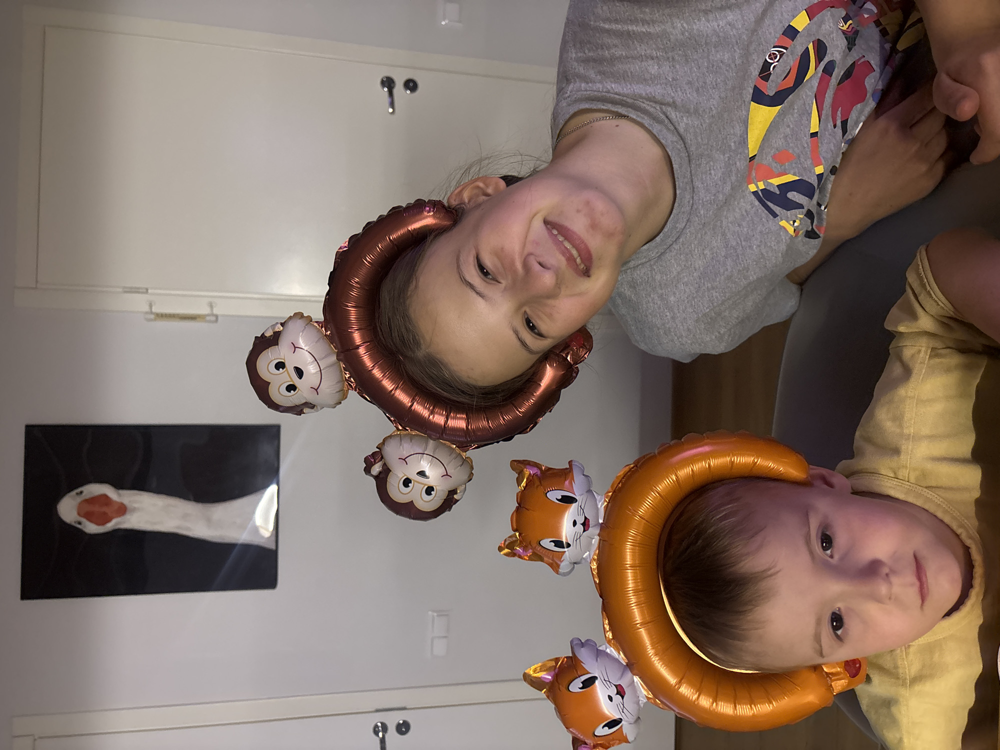 In a domestic indoor setting, two children, a boy and a girl, are posed with novelty animal-shaped balloons. The boy, positioned in the foreground on the left, wears an orange, ring-shaped balloon on his head, adorned with two fox-shaped balloons. He has light brown hair and is looking directly at the camera with a neutral expression. He is wearing a yellow shirt. The girl, on the right and slightly behind the boy, is wearing a large, copper-colored ring balloon on her head, with two monkey-shaped balloons attached. She has brown hair and is smiling at the camera. She is wearing a grey t-shirt with a colorful graphic design. In the background, on the wall, hangs a framed artwork of a goose's head against a black background. Two doors, one on the left and one on the right, are visible. The location is a residence in Chaikovsky, Russia.