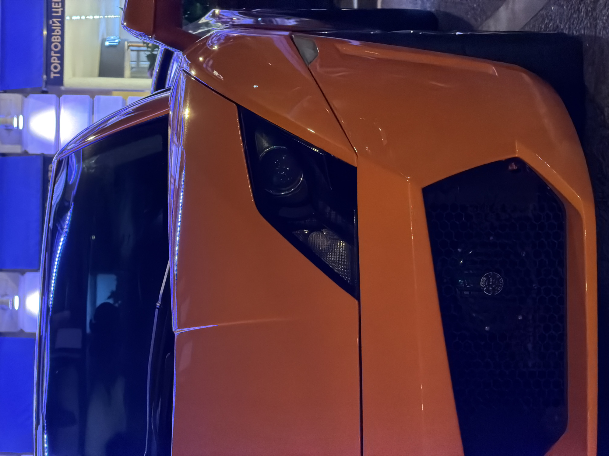 The image is a close-up of the front passenger side of an orange Lamborghini, possibly a Murciélago, taken at night. The car is parked on a city street. The headlight and front grille are prominently visible. In the background, there is a building with signage in Russian that translates to "Shopping Center". Based on the signage and the car model, the location is likely in Moscow, Russia. There are no people visible in the image. The focus is on the vehicle's exterior design and features. The environment is urban, with street lighting casting a glow on the car.