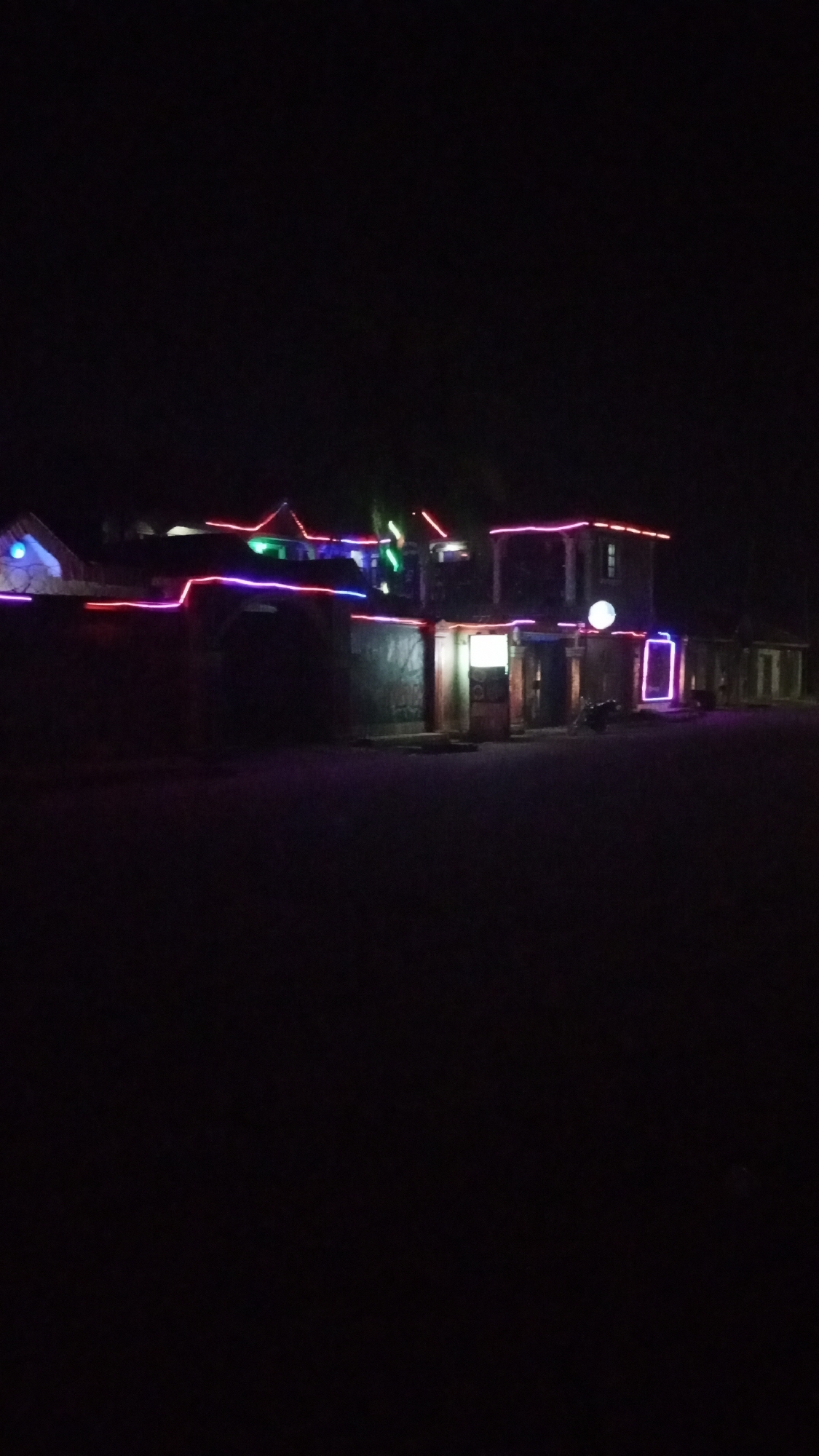 The image shows a dimly lit building in Sabon Tasha, Nigeria, at night. The building is decorated with strings of colorful lights along the roof and around doorways. A motorcycle is parked outside the building. The surrounding area is dark with minimal visible detail. No people are readily apparent in the image.