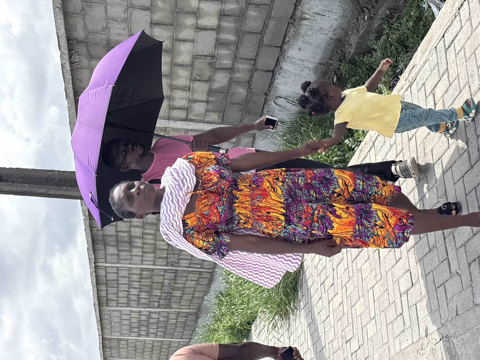 This image depicts a group of people walking outdoors on a paved pathway under an overcast sky. In the foreground, a woman is seen facing the viewer, wearing a vibrant, multi-colored patterned dress in hues of orange, yellow, purple, blue, and red, along with a white and purple striped scarf draped over her shoulders. Behind her, a man in a pink t-shirt holds a large purple and black umbrella above them, while also holding a black smartphone in his other hand. To the right, a small child, likely a girl with her hair styled in pigtails, walks in a bright yellow t-shirt, blue patterned pants, and colorful sneakers. Her attention is drawn away from the camera. The ground beneath them is composed of light-colored interlocking bricks, with green grass and bushes bordering the pathway on the right side. A large concrete structure, possibly part of an overhead bridge or building, is visible in the background under the cloudy sky. The scene suggests a casual daytime outing, possibly for protection from the sun or light rain, set in an urban or semi-urban environment in Igboefon, Nigeria. No visible text is present in the image.