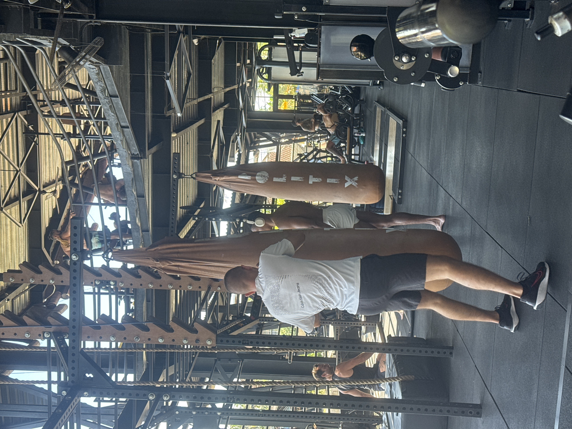 The image depicts an indoor gym setting in the Ubud District, Indonesia, characterized by its industrial-chic aesthetic and natural light. In the foreground, a man with a shaved head wears a white t-shirt with "Calvin Klein Jeans" printed on the back and dark shorts. He is positioned facing away from the camera, in front of a large, tan punching bag. To his right, another man, partially visible, wears white shorts and headphones. To the left, another man in a dark tank top is seated on a dark box.
The gym's structure is a prominent feature, with exposed metal beams, wooden accents, and a corrugated metal roof visible overhead. Several punching bags hang from the ceiling, one of which has "LITIX" printed vertically. In the background, multiple levels of platforms and structures are present, populated by other gym-goers engaged in various activities. One individual is seen lying on a bench and lifting weights, while others are visible on elevated platforms. Gym equipment, including weight machines and racks, are scattered throughout the space. Natural light streams in from windows, illuminating the scene and casting shadows. The flooring is a dark rubber mat.