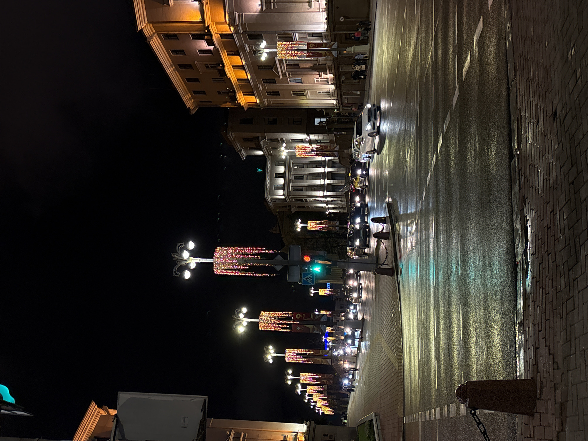 Captured at night in Minsk, Belarus, the image shows a well-lit urban street scene. A row of ornate street lamps, decorated with string lights, lines the sidewalk. The street is wet, reflecting the lights from the street lamps and passing vehicles. Buildings with illuminated facades stand on one side of the street, while darkness fills the other side of the frame. Cars are parked along the curb, and there are a few pedestrians visible on the sidewalk. A traffic light is showing a green light.