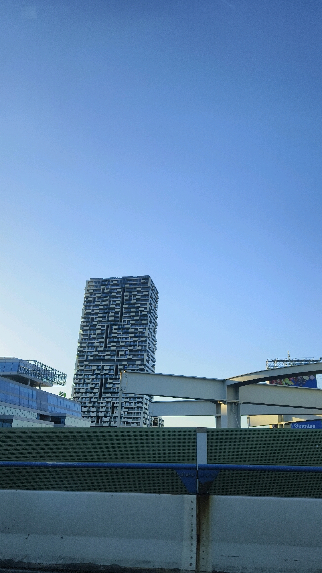 The image shows a view of the Danube area in Vienna, Austria, from an elevated perspective, likely from a highway. In the foreground, there is a concrete barrier with a green mesh fence behind it, and a blue railing. Several large modern buildings are visible in the midground. The most prominent is a tall, slender skyscraper with a distinctive facade of dark balconies and white walls. To the left of this skyscraper, there are other modern buildings with glass exteriors. In the background, the sky is clear and blue, suggesting it is daytime. A large billboard is also visible in the background with the word "Gemüse" (German for "vegetables") on it. The scene appears to be an urban landscape, characterized by contemporary architecture and infrastructure.