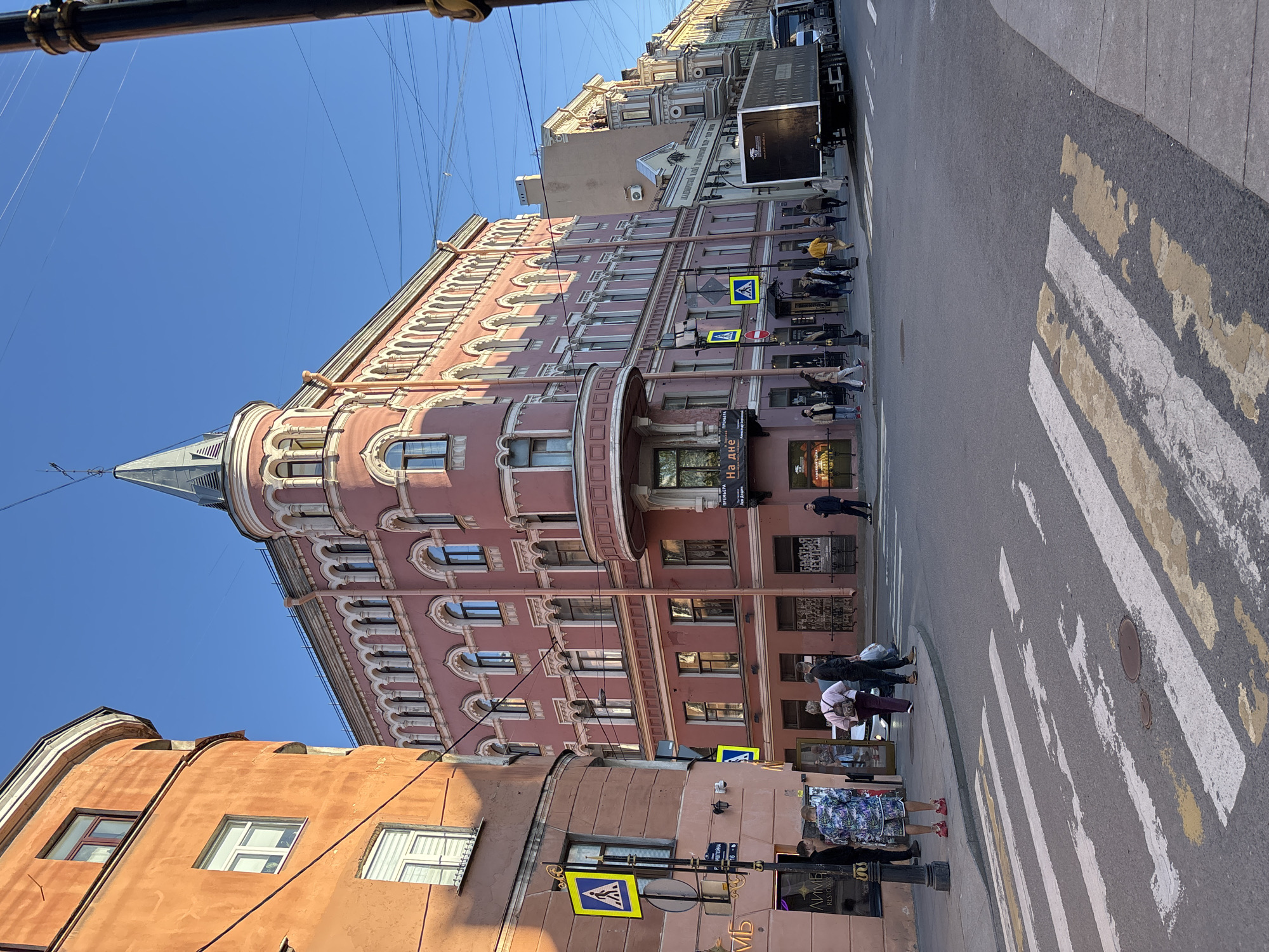 The image shows a street scene in Saint Petersburg, Russia. A multi-story pink building with architectural details like arches and a pointed tower occupies a prominent position. Pedestrians are waiting on the sidewalk near a zebra crossing. Street signs with pedestrian crossing symbols are visible. A truck is parked further down the street. The sky is clear and blue.