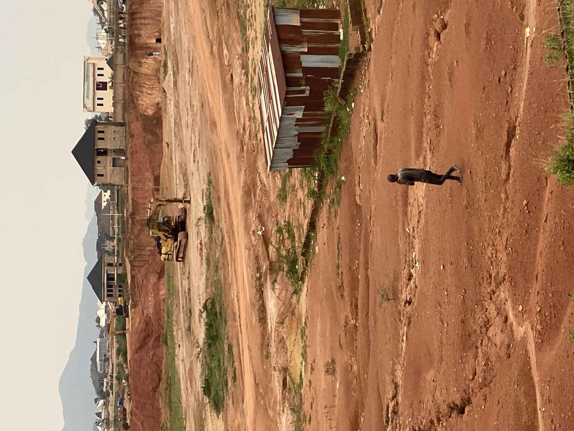 The image depicts an outdoor scene in the city of Jos, Nigeria, characterized by a construction site with red earth terrain. In the foreground, a lone man walks across the dusty ground, wearing a dark t-shirt and pants. Further into the image, a dilapidated corrugated metal structure stands to the right, exhibiting signs of rust and wear. In the middle ground, a yellow excavator is positioned on the uneven earth, suggesting ongoing construction or excavation work. In the background, several unfinished or newly constructed houses are visible, along with more excavators and a mountainous landscape under a cloudy sky. The overall impression is of a developing area with ongoing construction activities.