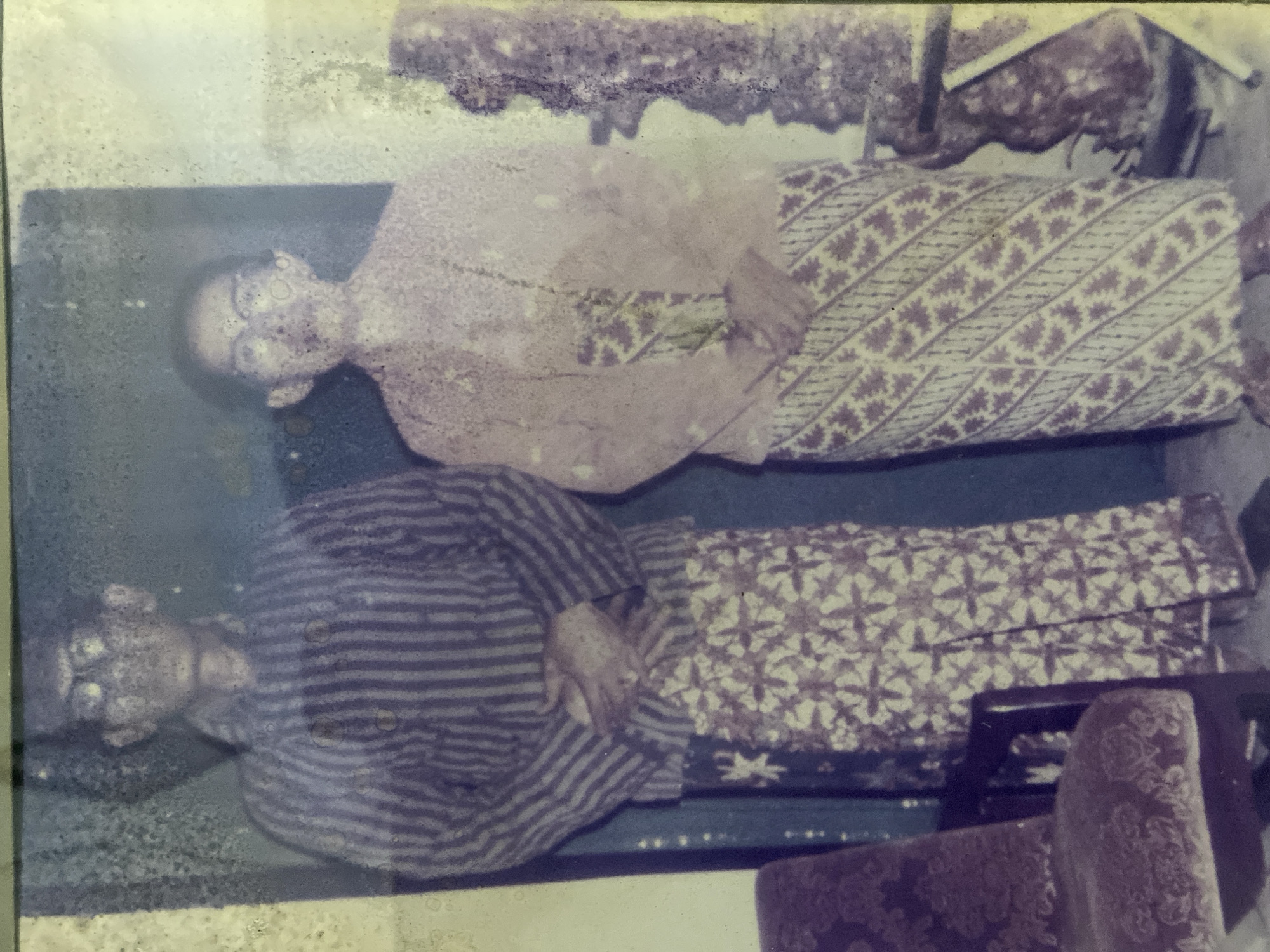 A faded, vintage, full-length studio portrait depicts a man and a woman, likely a couple, standing side-by-side. The man, on the left, wears a striped, collared shirt and a batik sarong with a repeating star pattern. His hands are clasped in front of him. The woman, on the right, wears a lighter colored, possibly silk, blouse and a batik sarong with a diagonal geometric pattern. She also has her hands clasped. Both individuals wear glasses. In the foreground, a dark-colored, upholstered chair with carved wooden arms is visible on the far left. The background appears to be a plain studio backdrop, with a hint of a dark structure on the right side of the frame. The overall image quality is degraded due to age, with visible fading, discoloration, and specks of dirt or damage on the photograph. The setting is not specifically identifiable as being within the city of Kediri City, Indonesia, as there are no distinct landmarks or environmental cues to confirm the location.