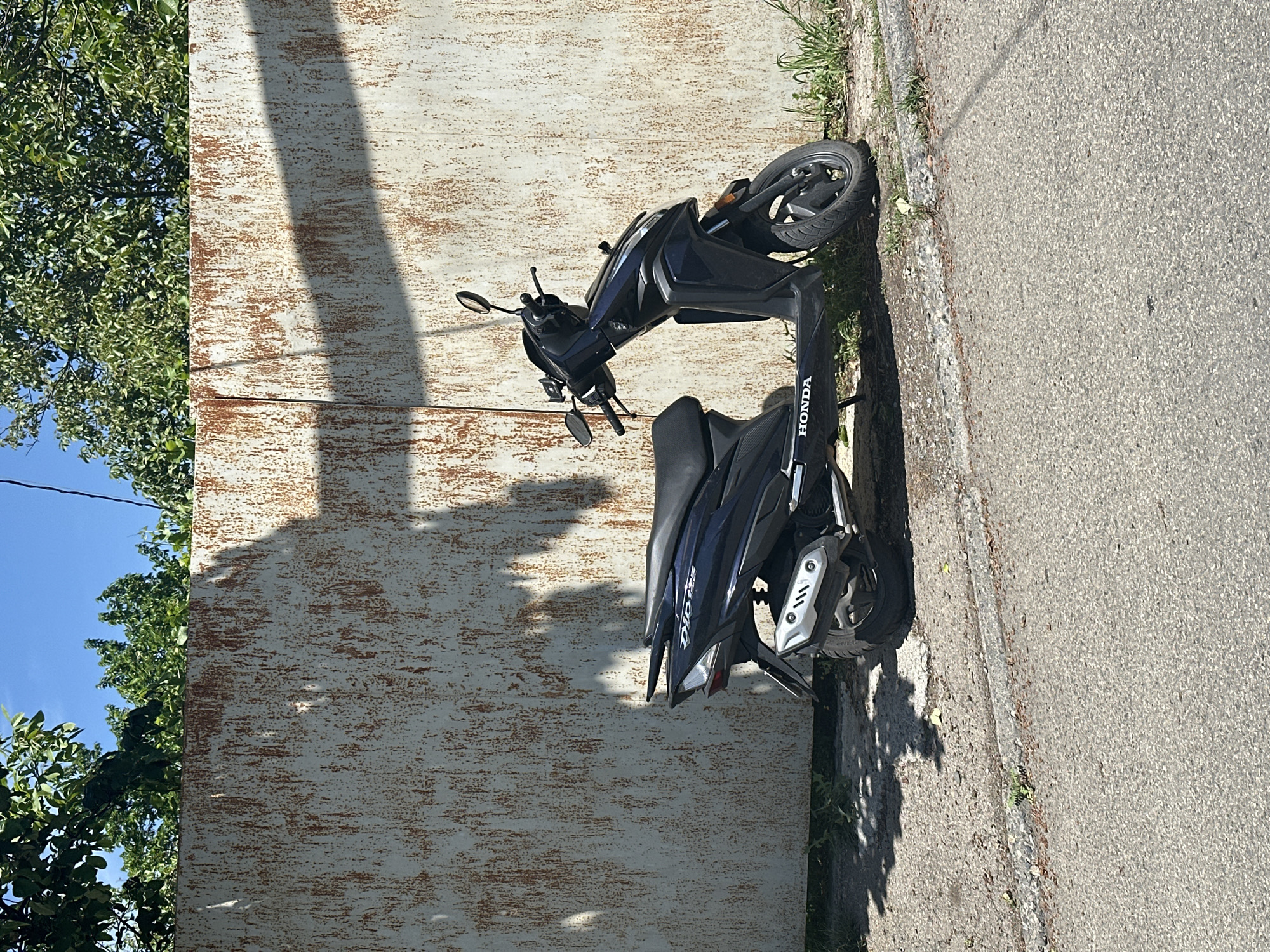 A blue Honda scooter is parked on the side of a road in Tbilisi, Georgia. The scooter is situated next to a weathered, white-painted metal wall. A tree with green foliage is partially visible to the left of the wall, with a blue sky above. The road is asphalt. The photograph is taken in daylight.