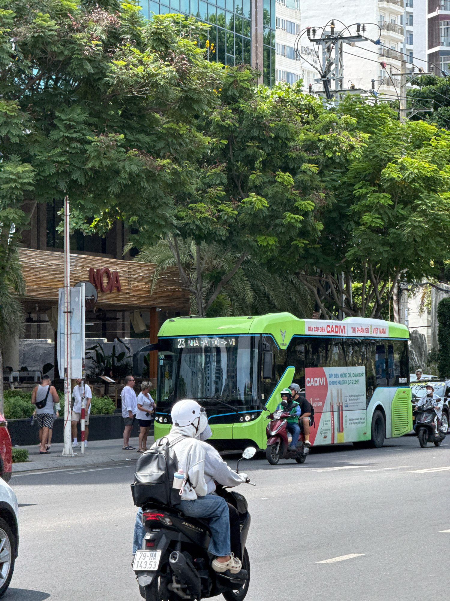 The image depicts a bustling street scene in Nha Trang, Vietnam, on a bright, sunny day. In the foreground, a person is riding a dark-colored scooter, wearing a light-colored long-sleeved shirt, blue jeans, a white helmet, and a black backpack. The scooter has a license plate reading "79-HA 143.53". To the right, a green and white bus with "CADIVI" branding is visible, with text in Vietnamese on its side. Two people are seated on a red scooter behind the bus, both wearing helmets. Further to the right, another scooter with two occupants, one wearing a white mask, is also present.
In the background, several people are standing on a sidewalk. Behind them is a building with a facade made of bamboo-like material, displaying the red sign "NOA". Beyond that, modern buildings with glass windows and balconies rise up, interspersed with lush green trees and power lines. Palm trees are also visible, suggesting a tropical or subtropical climate. The street is paved and appears to be a main thoroughfare. The overall atmosphere is that of an active urban environment.