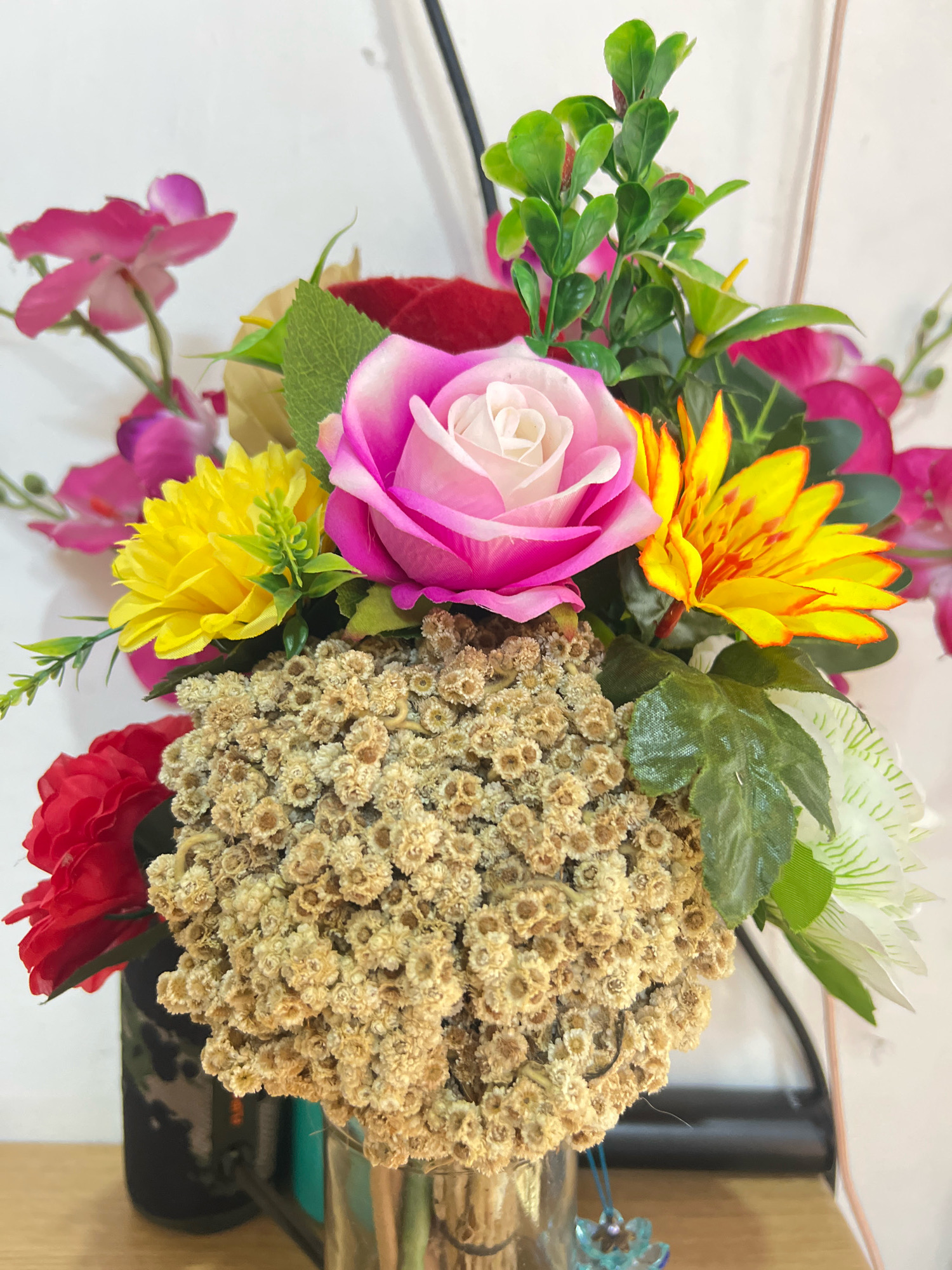 This is a close-up, eye-level, indoor photograph of a vibrant artificial flower arrangement. The bouquet features a prominent pink and white rose, a bright yellow daisy-like flower, and a striking yellow and orange flower with pointed petals, possibly a type of lily or lotus. Interspersed among these larger blooms are smaller, dried, straw-colored flowers that create a textured mass in the foreground. Accompanying these are red roses and delicate pink orchid-like flowers, along with various green artificial leaves. The flowers are arranged in a clear glass vase, which is partially visible at the bottom center. The background consists of a plain, light-colored wall, with a dark vertical object on the left and a blurred white object on the right, suggesting an indoor setting. The overall impression is one of a colorful and decorative display. The image does not provide any specific information about its location in Denpasar, Indonesia, or any individuals present.