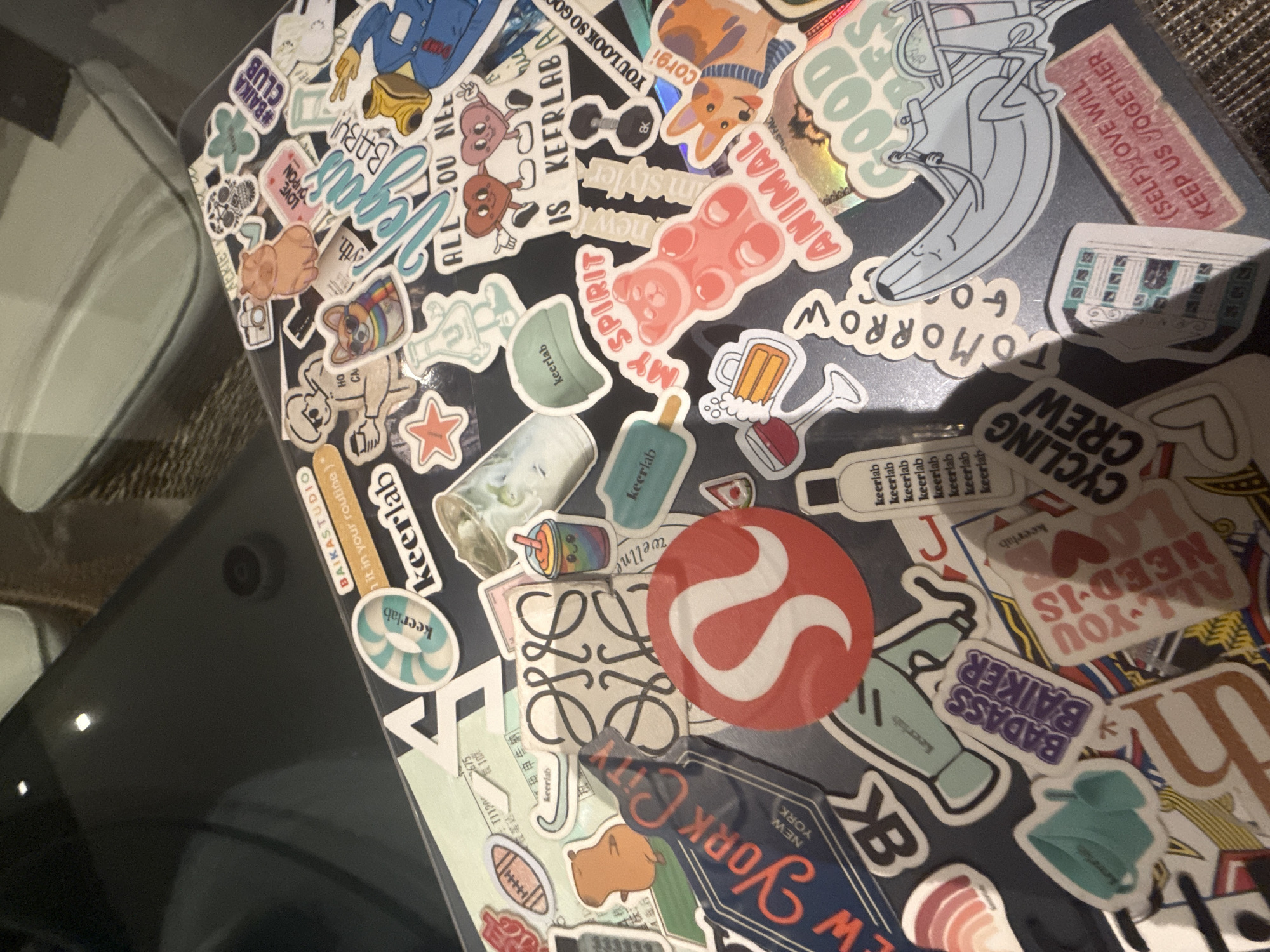 The image presents an angled, close-up view of a black laptop heavily covered with a dense array of colorful stickers. The laptop rests on a dark surface, with a blurred light-colored object or reflection visible on the left side of the frame.
The primary subject is the personalized laptop, distinguished by its extensive sticker collection. These stickers feature diverse designs, including brand logos such as the red Lululemon symbol, the interlocking 'L's of Loewe, and multiple "Keerlab" stickers, often in mint green depicting various items like hats, bottles, and popsicles. "Baika Studio" is also present.
Other stickers illustrate a variety of themes:
- **Characters and Animals:** A corgi wearing a hat, a pig-like figure, a pink gummy bear labeled "MY SPIRIT ANIMAL," a cartoon banana on a unicycle, and two heart-shaped characters.
- **Food and Drink:** A beer mug, a boba tea drink with a rainbow straw, a popsicle, and a ramen bowl.
- **Objects and Symbols:** A skull, a dumbbell, a camera, a football, a star, a clover, and various abstract patterns.
- **Text and Phrases:** Prominent text includes "ALL YOU NEED IS LOVE," "BADASS BAIKER," "CYCLING CREW," "YOU LOOK SO GOO," "(SELF)LOVE WILL KEEP US TOGETHER," "Vegas," "#BAIKA CLUB," "NEW YORK CITY," "hit in your routine," "MY SPIRIT ANIMAL," "new m styler," "LOVE COUPON," and "TOMORROW." The word "keerlab" appears frequently across many smaller stickers.
A small, circular camera lens is visible on the upper left side of the laptop's body. The lighting suggests an indoor setting. This highly customized laptop is located in San Bernardino Tlaxcalancingo, Mexico.