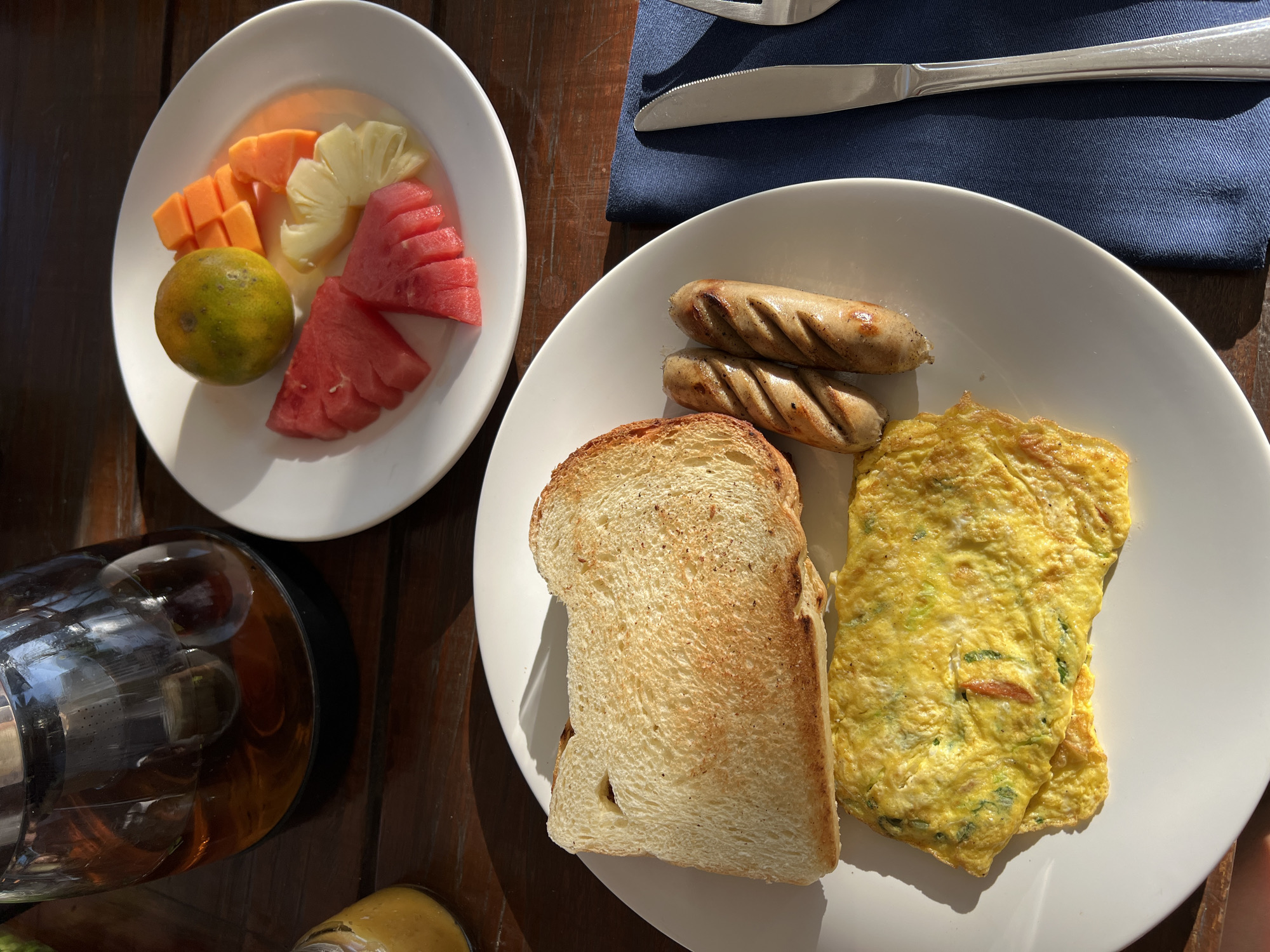 A breakfast spread is laid out on a wooden table. The meal includes a plate with a piece of toasted bread, an omelet with visible green specks, and two grilled sausages. Another plate features a variety of sliced fruits: watermelon, pineapple, papaya, and an orange. A glass teapot filled with amber liquid, likely tea, sits to the left of the fruit plate. A small jar containing a yellow substance, possibly jam or sauce, is also visible. A dark blue napkin with a fork and knife is positioned to the right of the main breakfast plate. The scene is illuminated by sunlight casting distinct shadows. This image was captured in Busungbiu, Indonesia.