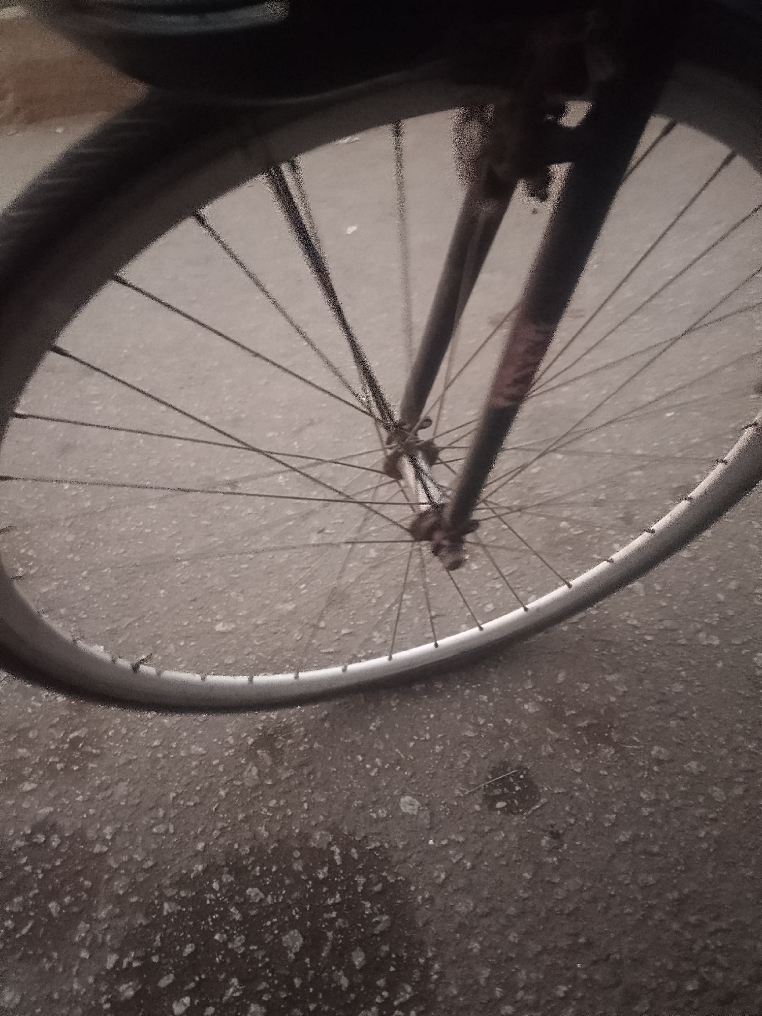 The image depicts the front wheel of a bicycle on a paved surface, potentially a road. The wheel has a silver rim and thin spokes. The tire is black. The bicycle fork, likely steel, is visible and appears worn with some rust. The ground is a dark color with a rough texture, suggesting asphalt or a similar material. The lack of detail makes it difficult to definitively identify the exact location in Bukuru, Nigeria. No people are visible in the frame, and the focus is solely on the bicycle wheel and the immediate surrounding surface.