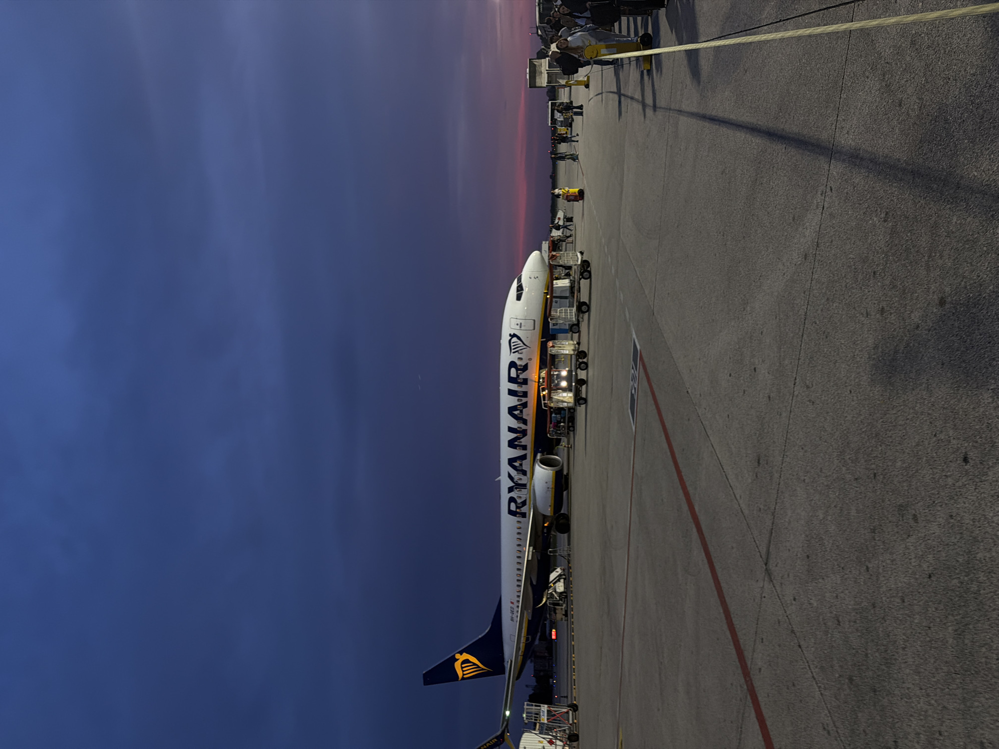 The image depicts a Ryanair Boeing 737 aircraft parked on the tarmac at an airport at dusk. The sky transitions from a deep blue to a reddish-orange horizon. Ground crew are positioned around the aircraft, with service vehicles alongside. A yellow rope stretches across the right side of the frame, seemingly delineating a work zone. The location is identifiable as an airport, possibly near Breda di Piave, Italy, based on the context.