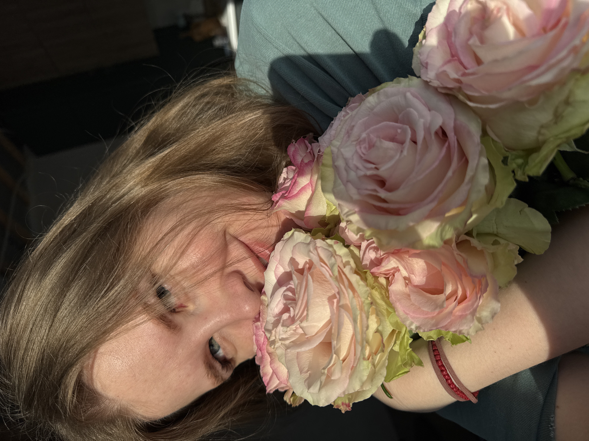 A blonde woman with blue eyes is holding a bouquet of light pink and white roses. She is wearing a teal shirt or dress and has a red bracelet on her wrist. The background is dark. The image appears to be taken in outdoor lighting. There is no information to place this image in the city of Gornyi.