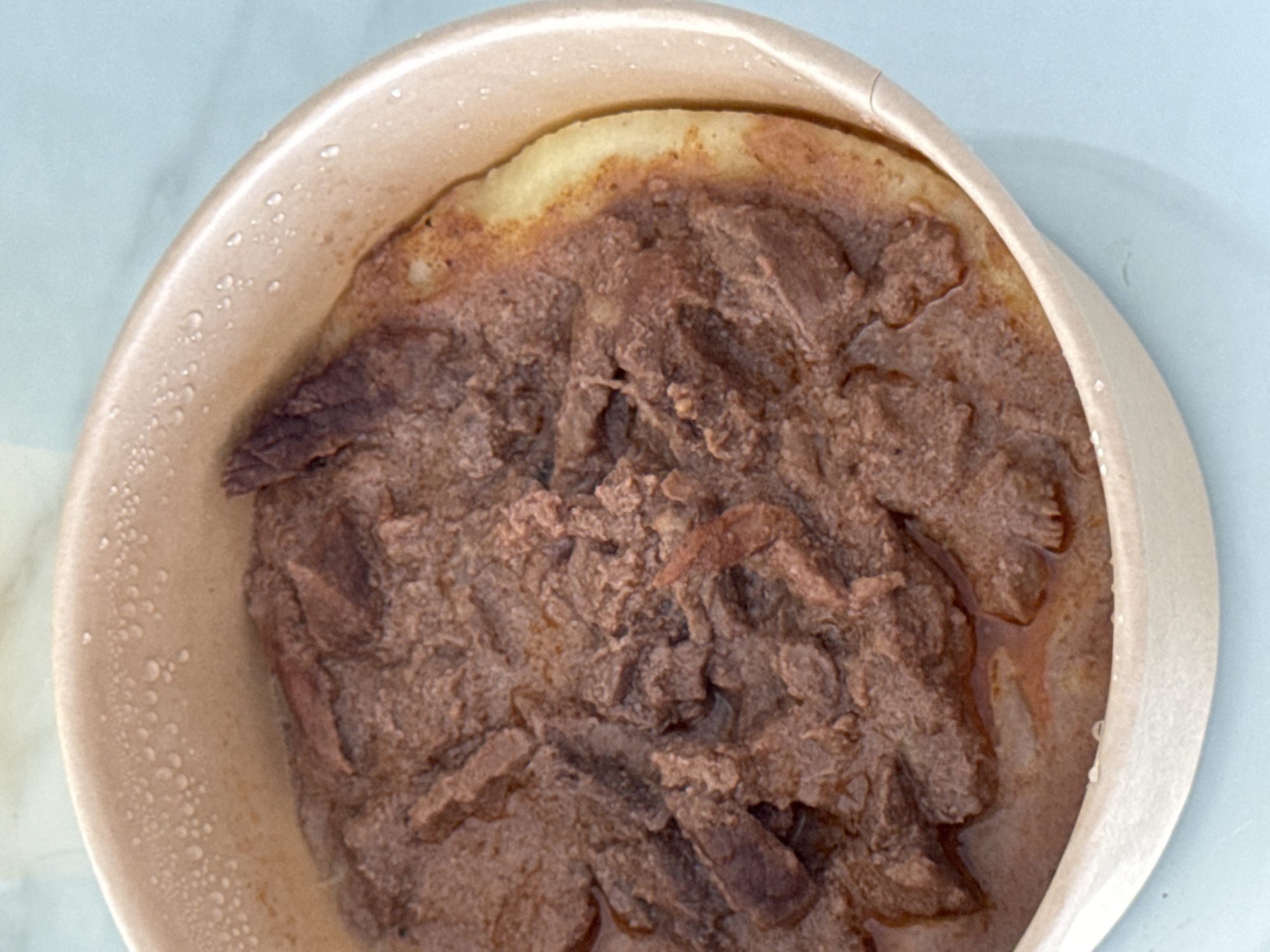 A close-up, overhead shot shows a dish of food in a light brown, disposable bowl. The food consists of shredded meat in a thick, reddish-brown sauce, layered over a pale yellow base that appears to be some type of bread or flatbread. The bowl has condensation droplets on its inner rim, and it is placed on a light blue surface. The scene is devoid of people and any discernible Bac Nha Trang Ward, Vietnam landmarks or environmental features.