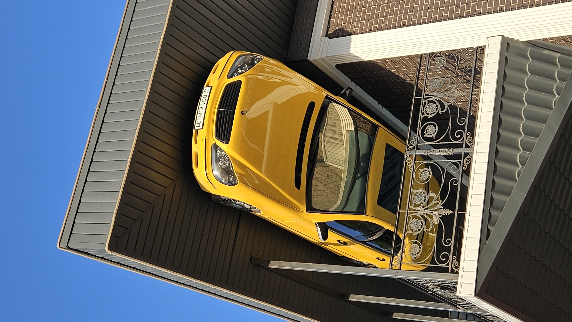The image depicts a yellow car precariously positioned on the roof of a building, a unique architectural display. The car is angled such that its front is elevated and its rear is lower, appearing as if it has been placed there intentionally. The building has a dark-colored roof with visible tile patterns, and a section of a brick wall is visible on the right side, adorned with a white vertical pillar. A balcony with an ornate metal railing separates the roof area from the lower part of the building. The sky above is clear and blue. The license plate of the car is partially visible and reads "001 KOK 01" with "KZ" indicating Kazakhstan. This unusual display is located in Almaty, Kazakhstan, specifically at the building of the "CAR BOUTIQUE" dealership.
