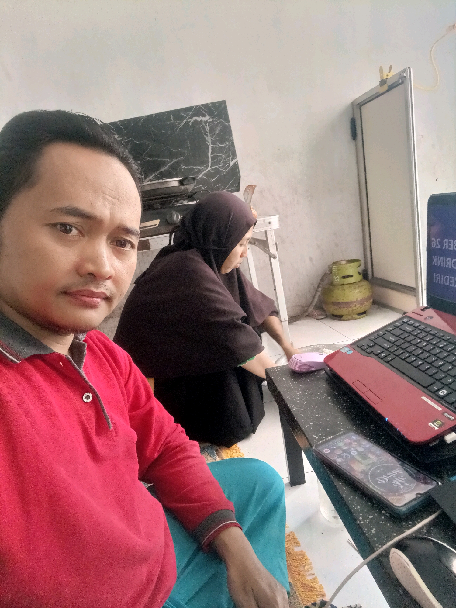 The image depicts an indoor scene, likely a home, in Badas, Indonesia. In the foreground, a man wearing a red collared shirt is taking a selfie. He has short black hair and is looking directly at the camera with a neutral expression. In the background, a woman wearing a dark hijab is kneeling and cleaning the floor. She appears to be using a cloth or sponge.
On a black countertop to the right, a red laptop is open, with its screen displaying text in a foreign language. A smartphone lies on the counter next to the laptop. A green gas cylinder is visible in the middle ground, to the left of the woman. The walls appear to be white, with some black marble-patterned tiles in the background. The overall environment suggests a casual, domestic setting.