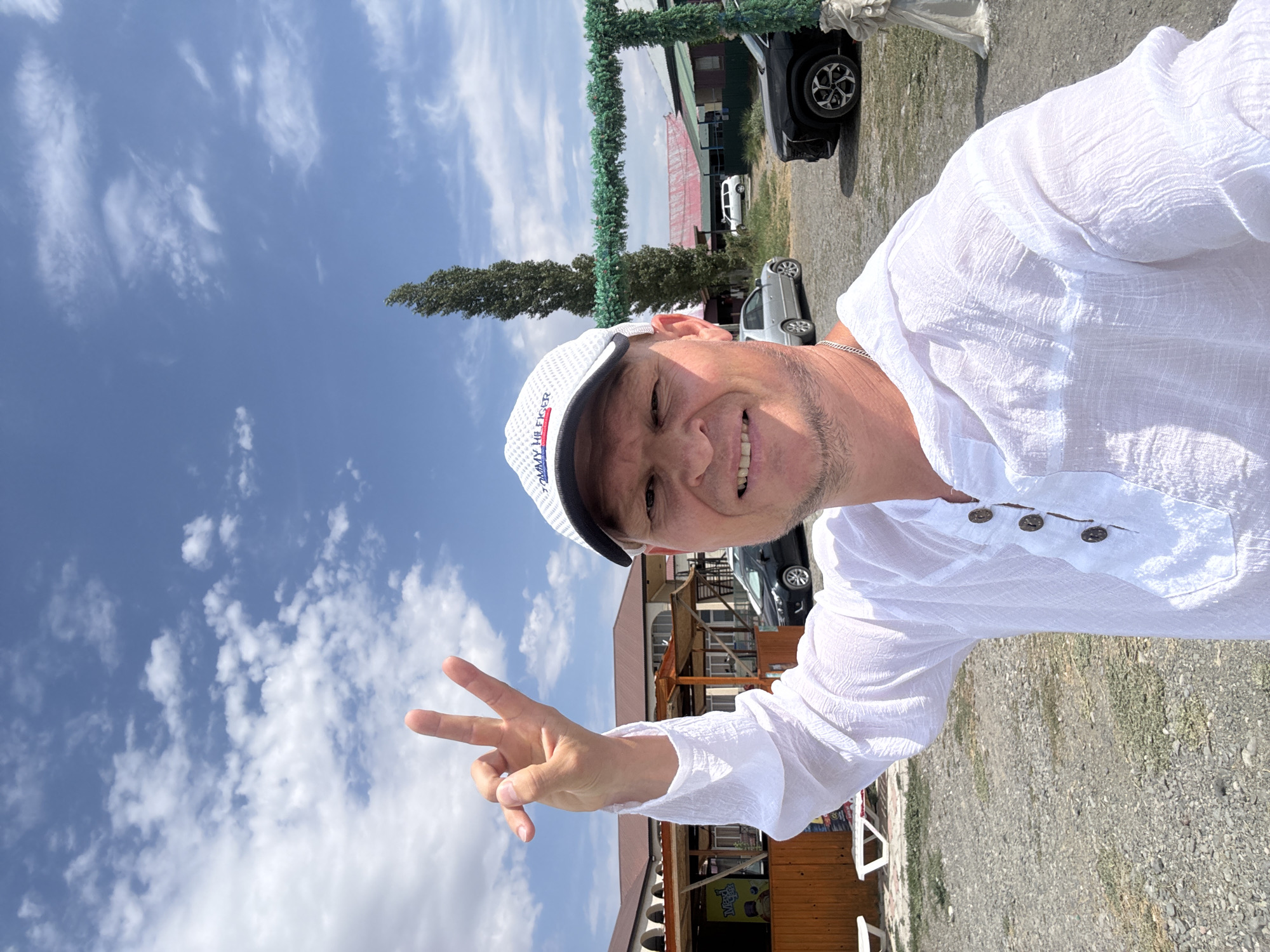 A man wearing a white Tommy Hilfiger baseball cap and a white linen shirt is taking a selfie outdoors. He is smiling and holding up his hand in a peace sign. In the background, there are buildings with brown roofs, a tall, slender tree, and several parked cars. The ground is covered in gravel. The sky is blue with scattered white clouds. The scene appears to be in a somewhat rural or semi-developed area, possibly a resort or roadside establishment. The location is identified as Urdzhar, Kazakhstan.
