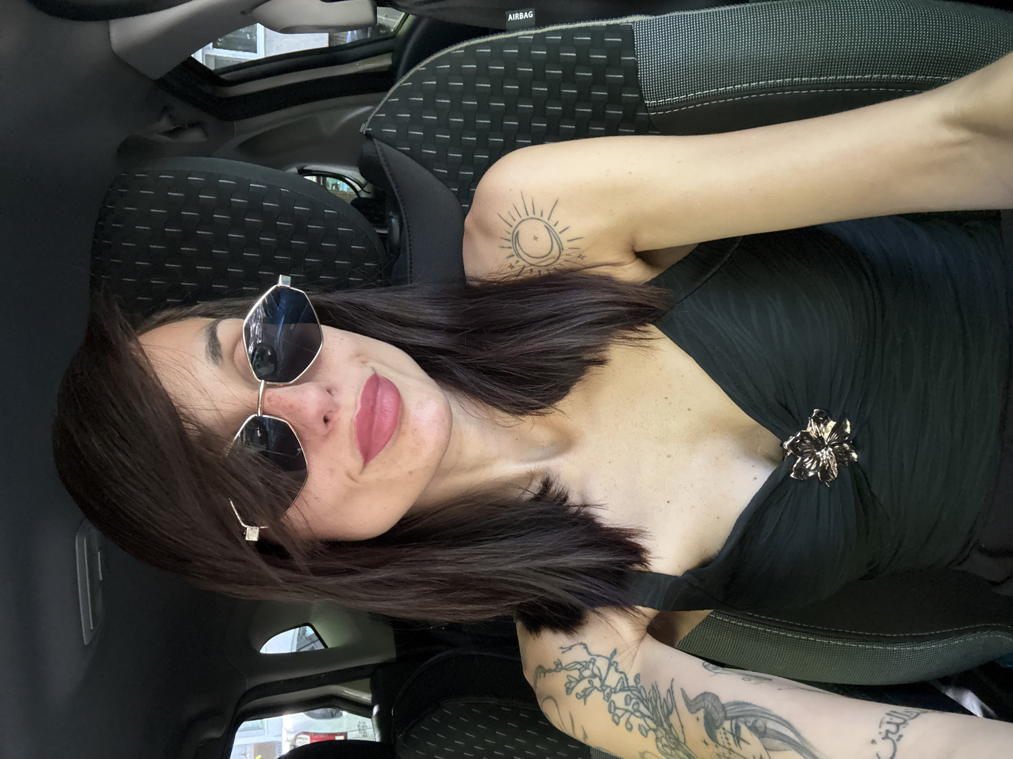 A woman is in a car. She has shoulder-length dark hair and is wearing sunglasses and a black top. There is a gold flower-shaped broach on the top. The woman has tattoos on her upper arms, one with a sun and moon design, and the other with a floral design. The car has dark grey seats and a black interior. The location is in Pleven, Bulgaria.