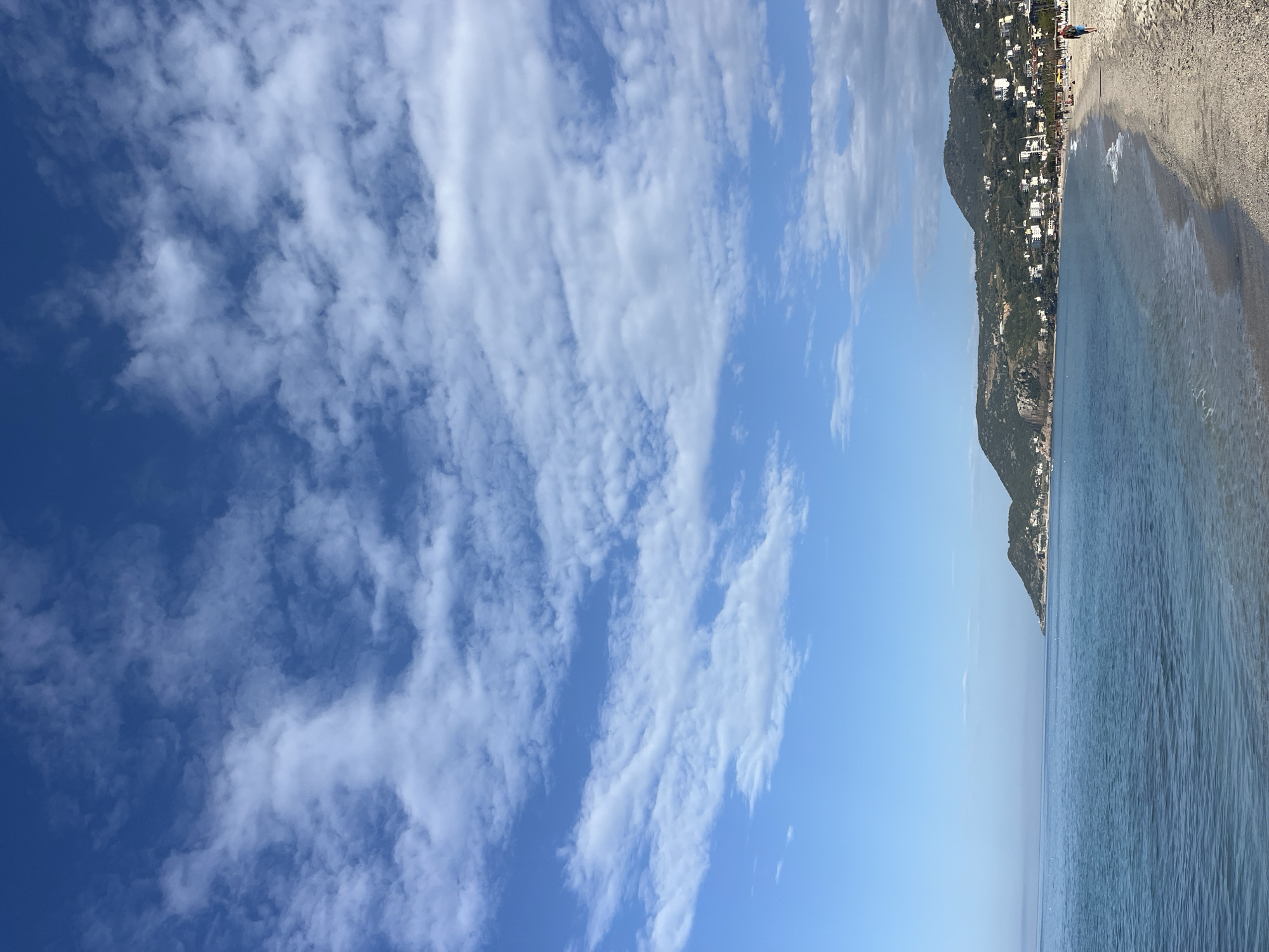 The image shows a beach scene in Alanya, Turkey. The sky is blue with wispy white clouds. To the right, a beach stretches along the coast, lined with buildings and vegetation that extends up a hill. The water is light blue near the shore, transitioning to a deeper blue further out. There are people on the beach.