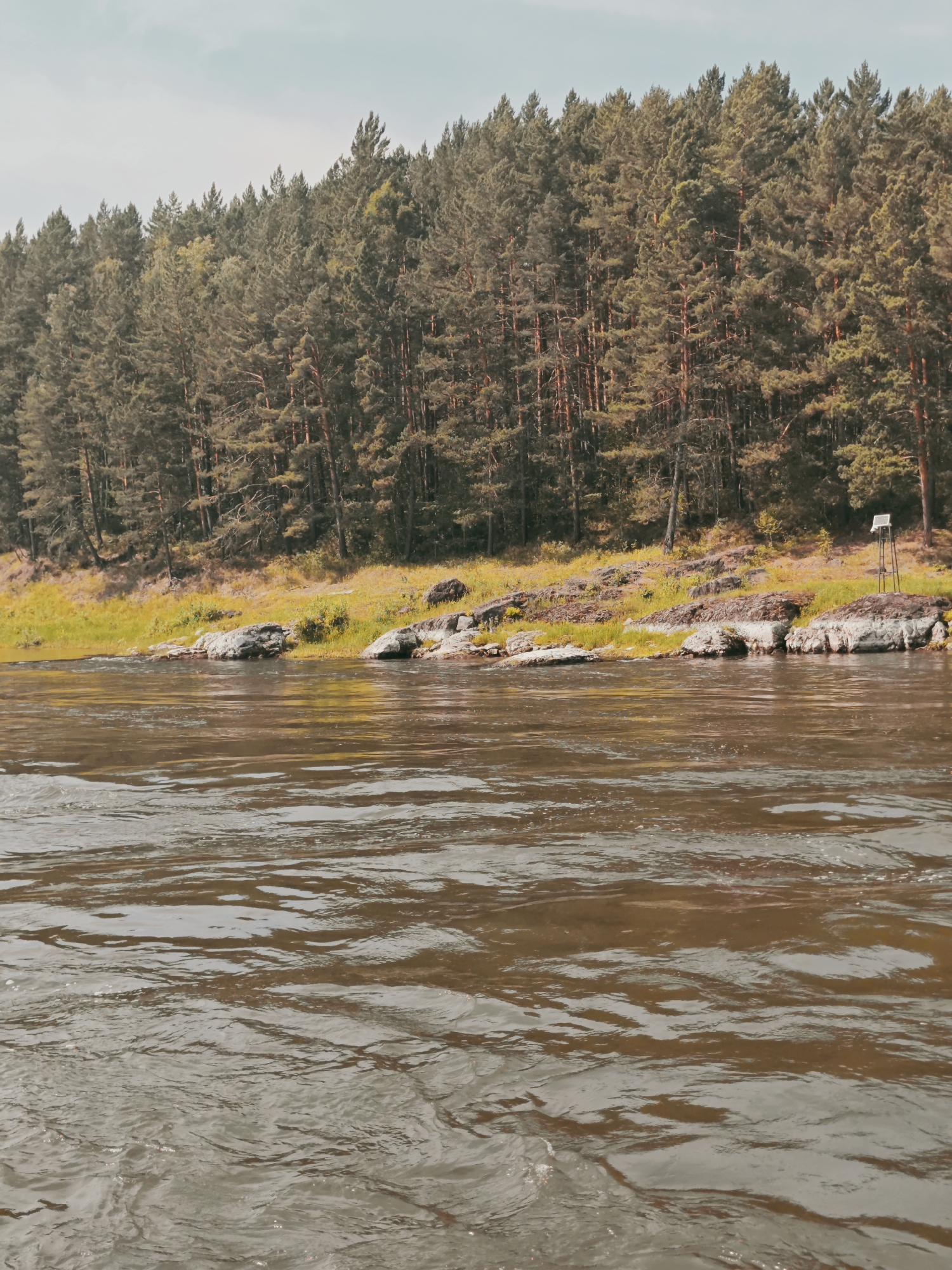The image depicts a serene natural landscape along the bank of a wide, brown river. The river occupies the foreground, its surface rippled and reflecting the light. The middle ground features a rocky, vegetated shoreline with scattered boulders. Beyond the shoreline, a dense forest of coniferous trees stretches across the horizon. The sky above is overcast with a pale, muted hue. There are no people or animals visible in the scene. A solitary, dark, weathered wooden post with a small solar panel on top stands on a rocky outcrop to the right side of the image, a subtle indication of human presence or monitoring equipment in this otherwise untouched natural environment. The location is identified as Porog, Russia, suggesting this is a view of the river in or near that city.