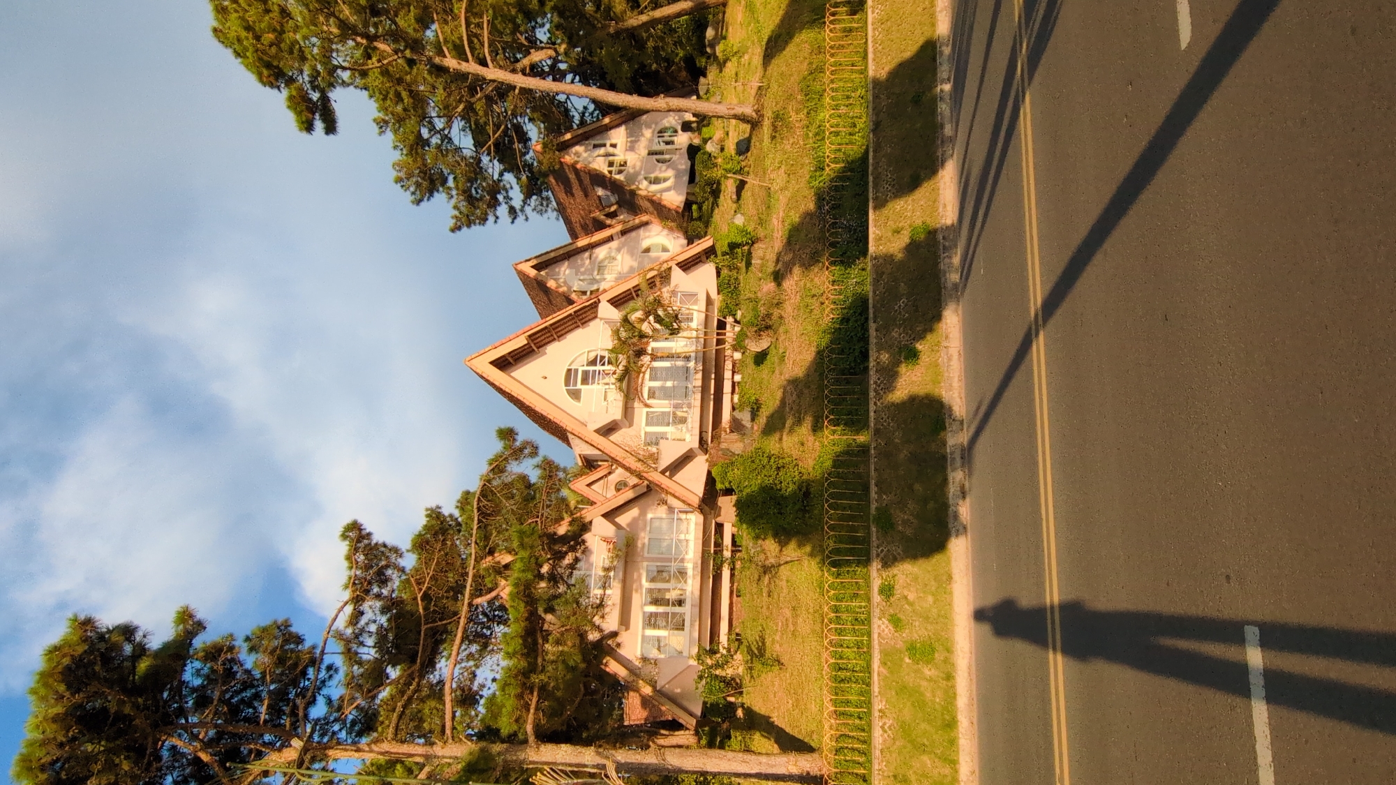 The image shows a row of houses with distinctive triangular roofs on a grassy hill. The houses are painted a light pink and have many windows. Trees, some with needle-like leaves, surround the houses and cast long shadows. In the foreground, a road runs horizontally, with a long shadow of a person taking the photo stretching across it. This scene is located on a street in Da Lat, Vietnam, known for its unique architecture and hilly terrain. The sky above is blue with scattered white clouds, and the lighting suggests it is late afternoon or early evening.