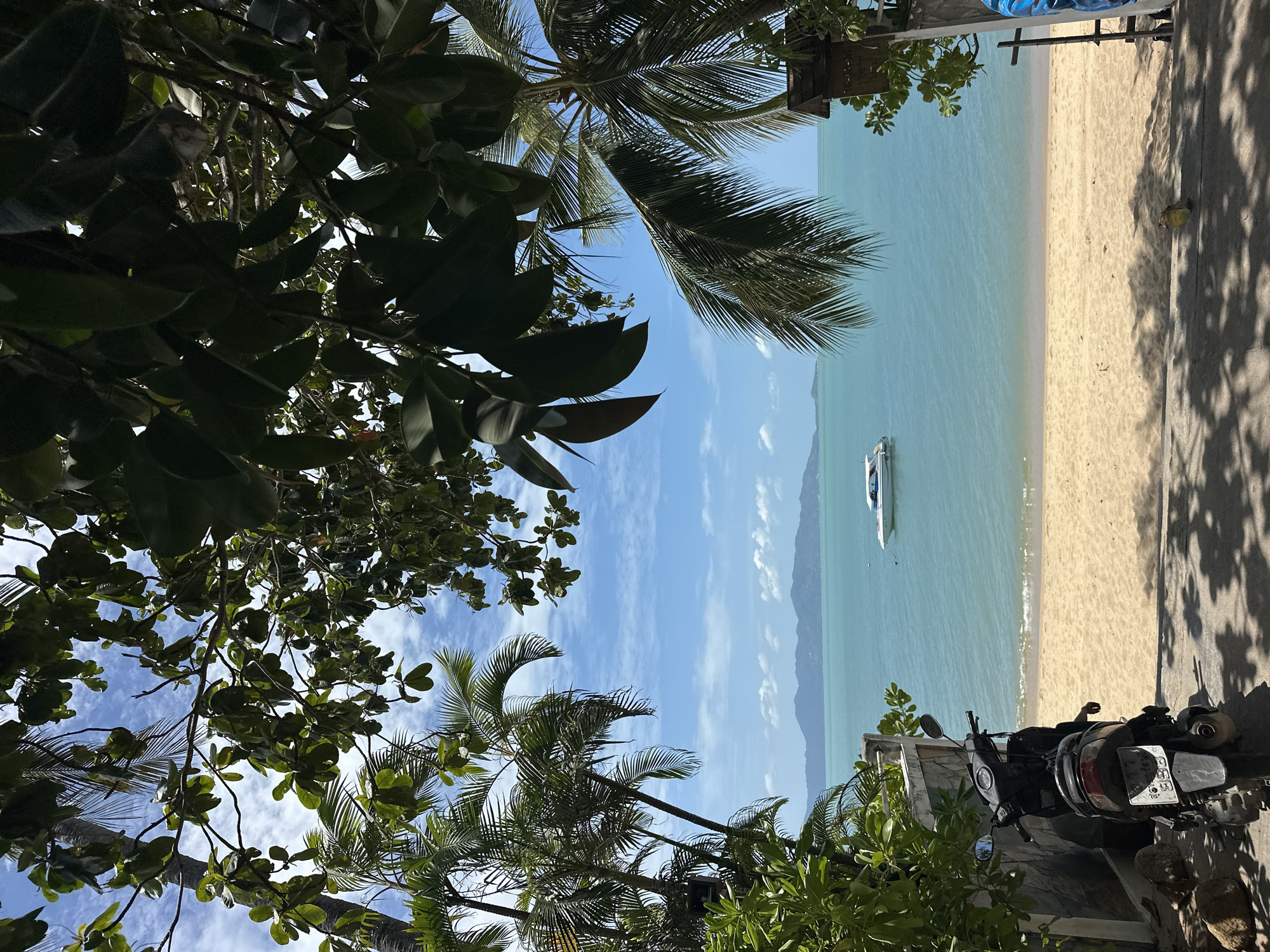 This is a medium-shot, eye-level, outdoor, daylight photograph taken in Koh Samui District, Thailand, looking out towards the ocean. The foreground features a dark gray scooter parked on a paved surface with shadows of palm fronds cast across it. To the left of the scooter, a stone wall borders a lush green bush.
The middle ground transitions to a sandy beach with a gentle slope leading down to the turquoise water. The water is calm with a small white boat visible in the distance, anchored offshore. Beyond the water, a hazy mountain range stretches across the horizon, meeting a bright blue sky dotted with scattered white clouds.
The scene is framed by abundant tropical foliage, with palm trees and dark green leaves creating a natural archway. The overall impression is one of a tranquil beachside setting.