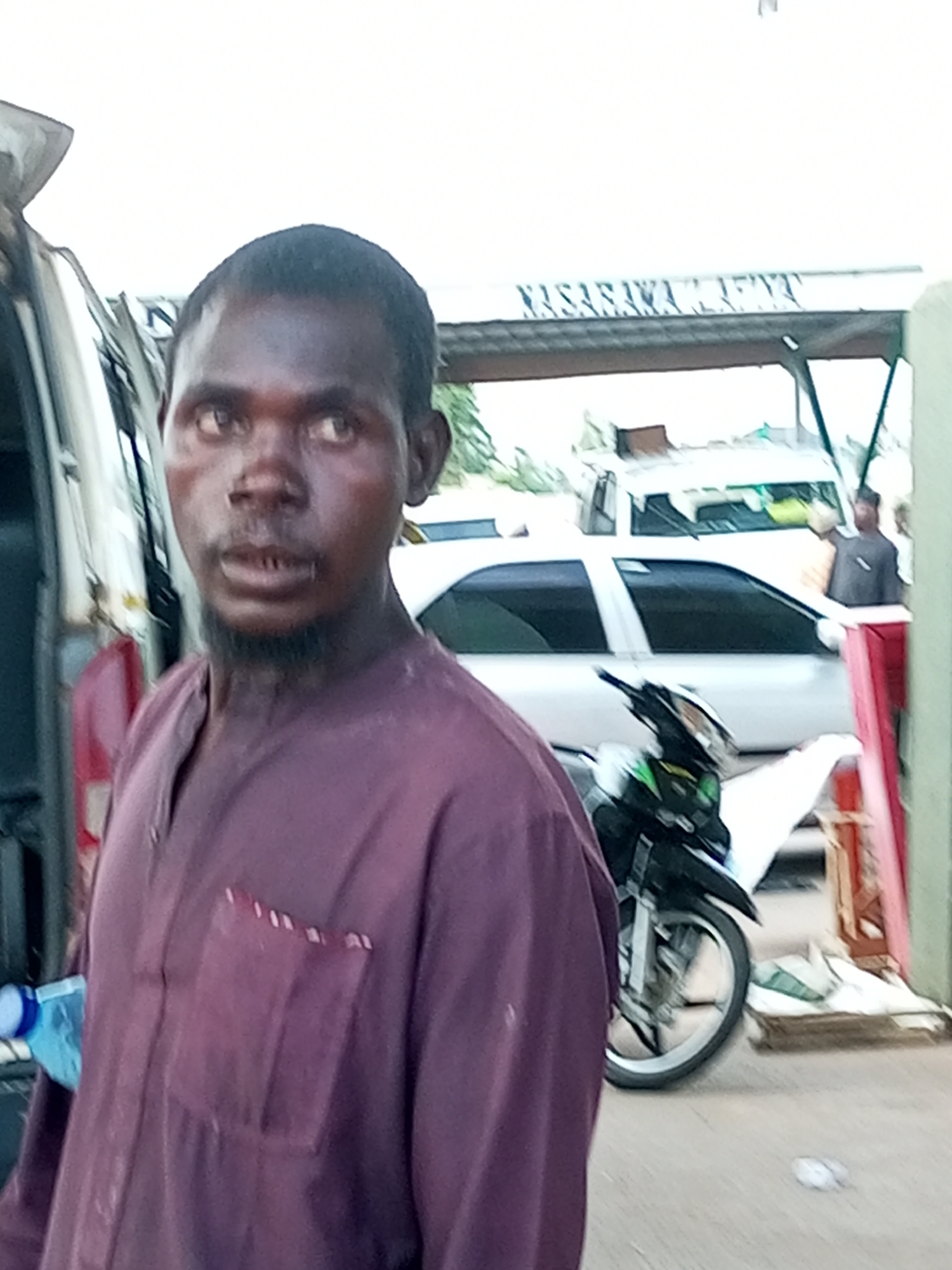 The image, taken in Gombe, Nigeria, depicts a man in a maroon garment standing in front of a white van. He has dark skin and short, dark hair, with a faint beard. His eyes are open, and he is looking slightly to his right. The van's door is open, and a blue and white object, possibly a water bottle, is visible near the bottom left. In the background, a white car is parked, and a black motorcycle is positioned to the right of the man. A sign with white lettering on a dark background is visible above the car, which appears to read "NASARAWA". The overall environment suggests an outdoor setting, possibly a street or parking area, with a clear sky overhead.
