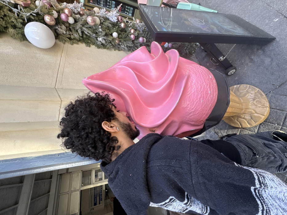 A person with curly dark hair appears to be kissing a large, pink, swirled, decorative ice cream cone. The individual is wearing a black hooded sweatshirt with white trim. A digital information screen stands near the ice cream. Holiday decorations, including green garlands, white lights, and pink ornaments adorn the wall behind the cone. The scene is captured in an outdoor setting, possibly near a building entrance.
