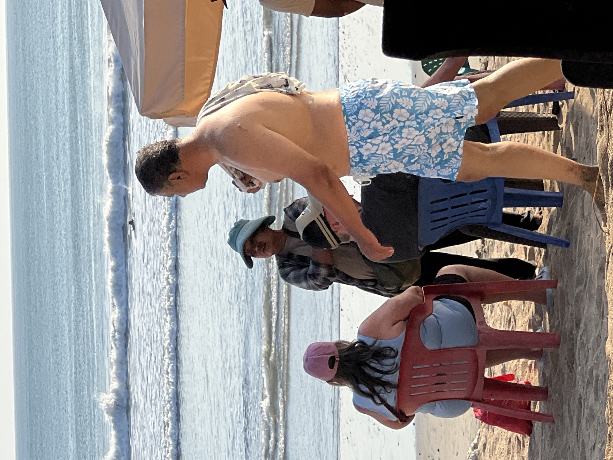 On a beach in Kuta, Indonesia, four individuals are positioned around beach chairs. A man, shirtless and wearing floral swim trunks, stands facing right. To his left, a person in a wide-brimmed hat and a plaid shirt is seated. In the foreground, a woman wearing a pink baseball cap and a light blue top is seated in a red chair. Another person is partially visible, seated in a blue chair to the right of the person in the plaid shirt. The ocean with gentle waves is visible in the background, and a tan canopy is set up on the right side of the frame. The scene appears to be a casual gathering on the beach.