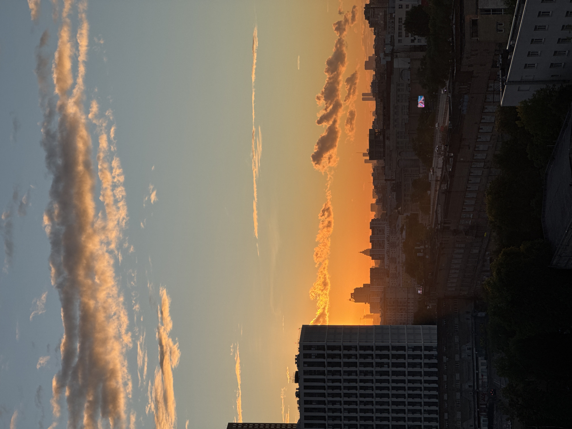 This is a vertical, medium-shot photograph of a city skyline at sunset. The sky is divided into two distinct color zones: the left side is a pale blue transitioning into a soft yellow, while the right side is dominated by vibrant orange and pink hues from the setting sun. Streaky, wispy clouds are visible throughout the sky, catching the warm light of the sunset.
In the foreground, a large, modern building with a grid-like pattern of windows takes up a significant portion of the lower frame. Behind it, the cityscape unfolds, characterized by a dense collection of buildings of varying heights and architectural styles. Some appear to be older, while others are more contemporary. Trees are interspersed among the buildings, appearing dark and lush against the illuminated sky. The overall scene depicts an urban environment as daylight fades, casting a dramatic and colorful light over the city.
The time of day is clearly sunset, indicated by the intense orange and pink colors in the sky and the long shadows cast by the buildings. The weather appears clear, with the clouds being a feature of the sunset rather than an indication of inclement conditions. The presence of distinct architectural styles and the mention of Moscow, Russia, suggest an urban setting within that location. There is an emotional tone of warmth and beauty conveyed by the sunset, contrasted with the more somber and imposing presence of the buildings.
A notable detail is a digital billboard located on a building further back in the cityscape, displaying a colorful advertisement or image. There are no people explicitly visible in the image, but the presence of vehicles on a road in the distance implies ongoing city life. The photograph captures a moment of transition from day to night in a bustling metropolitan area, highlighted by the dramatic natural light display.