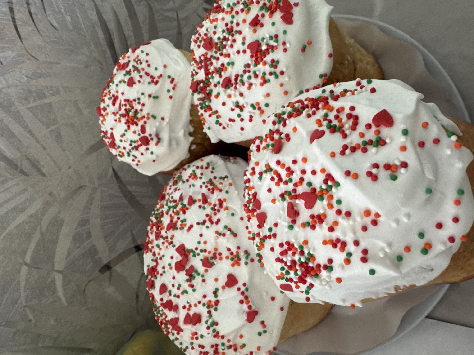 The image shows four pastries arranged in a light-colored bowl. Each pastry has a rounded top covered with white icing and decorated with red heart-shaped sprinkles, along with red, green, and white round sprinkles. The pastries appear to be close together, filling the bowl. The background is a soft grey color with what looks like a textured pattern on one side. The location in the city of Chaikovsky, Russia, is not discernible from the image.
