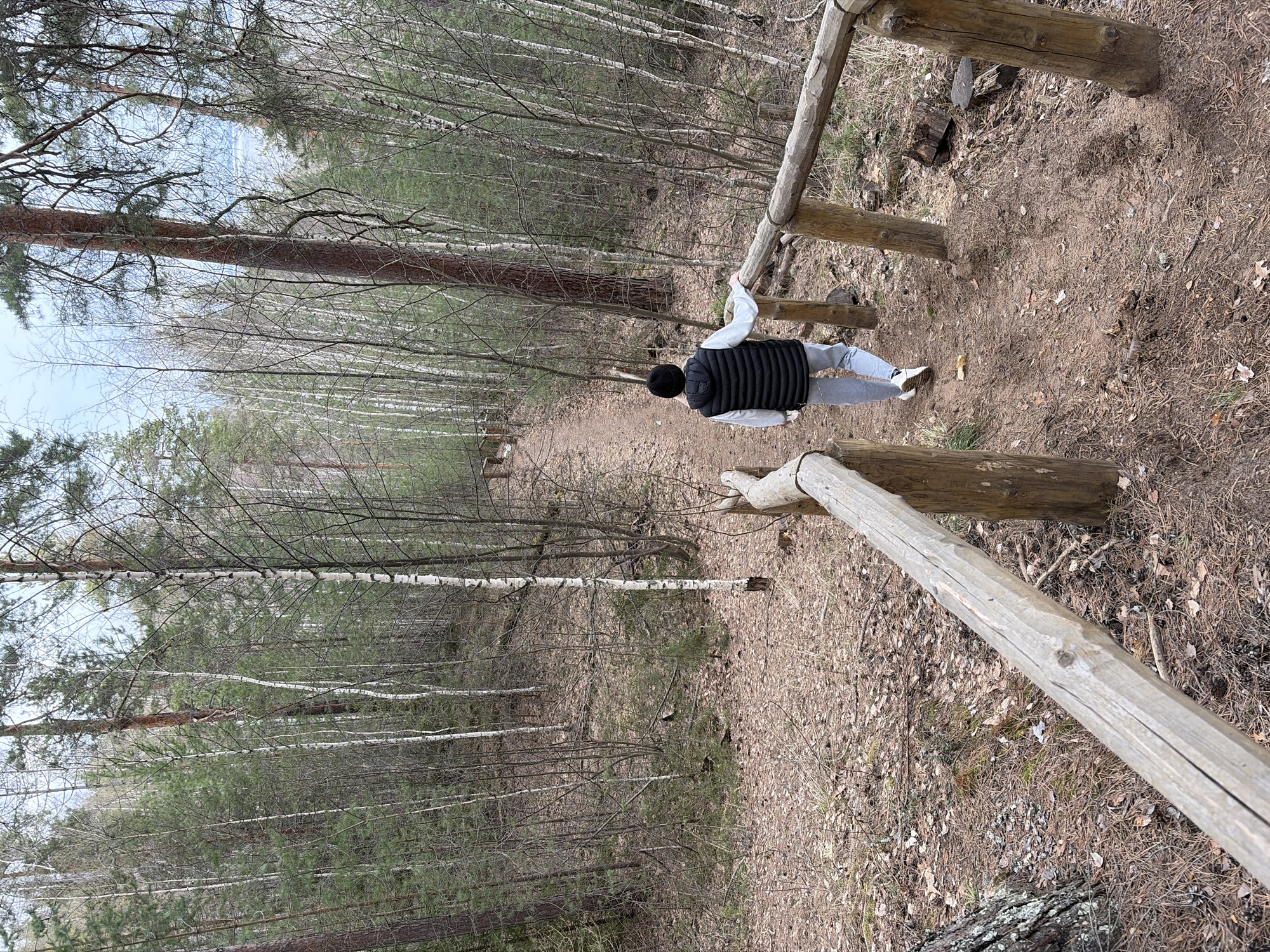 The image depicts a person standing on a path within a wooded area in Chaikovsky, Russia. The path descends, and a rough wooden railing is present on the right. The person is wearing a black vest, light gray pants, a light gray jacket, and white sneakers, and has a black beanie. The trees are mostly bare, suggesting a time of year before foliage. The ground is covered with leaf litter and pine needles.