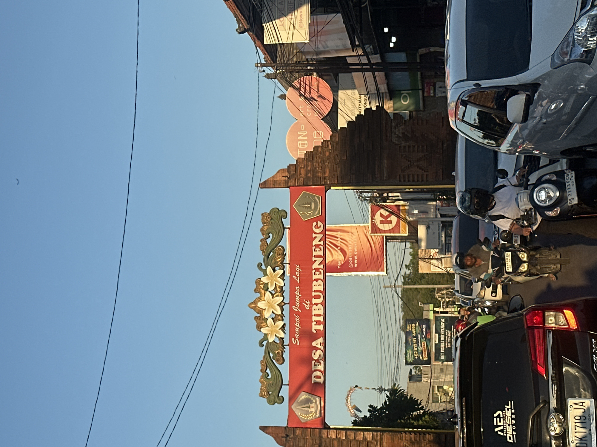 The image captures a street scene in North Kuta, Indonesia, with a traditional Balinese archway prominently featured. The arch, adorned with floral motifs and intricate carvings, bears the inscription "DESA TIBUBENENG" in large, bold letters. Beneath this, smaller text reads "Sampai Jumpa Lagi di" which translates to "See you again in".
The archway frames a busy street with vehicles, including cars and motorcycles, navigating through traffic. Several individuals are visible on the motorcycles, some wearing helmets. Billboards and signs with Indonesian text are placed along the roadside, indicating commercial activity in the area. The sky above is clear and blue, suggesting it is daytime. The overall impression is one of a lively and active urban environment in Bali.