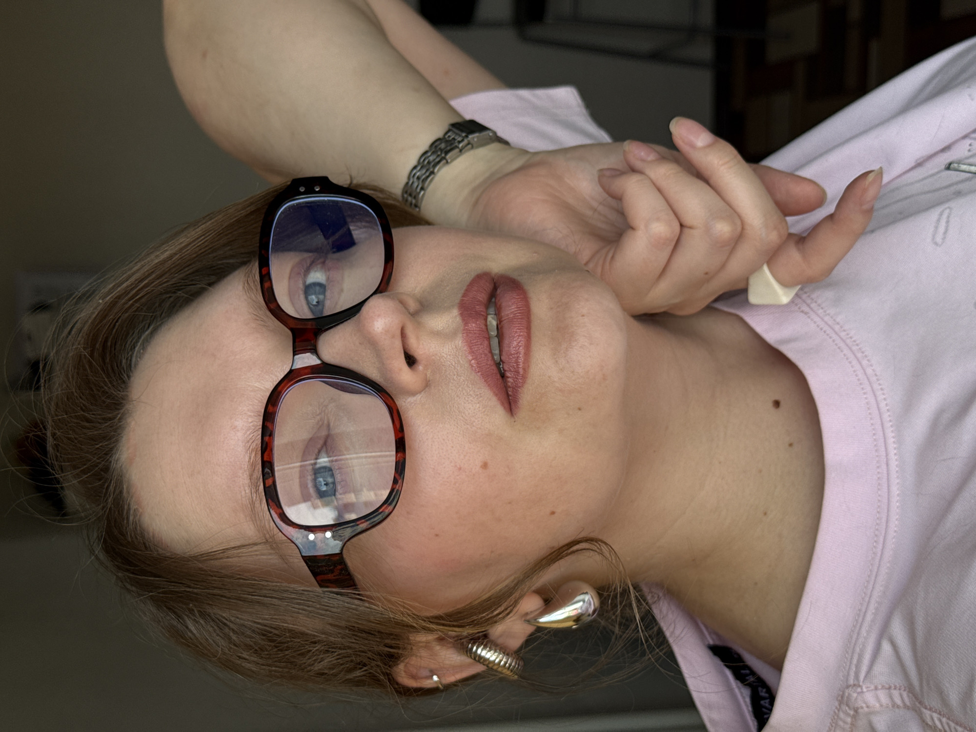 A woman lies at an odd angle, with her head angled downwards and to the left. She wears glasses with a dark brown, mottled frame. Her lipstick is a muted rose color. She wears a silver wristwatch on her left wrist and a large, off-white ring on her left hand. She has two earrings in her left ear, one a large, teardrop-shaped silver earring and the other a small silver hoop. She wears a pink shirt. The city of Izhevsk, Russia, is not present in the image.