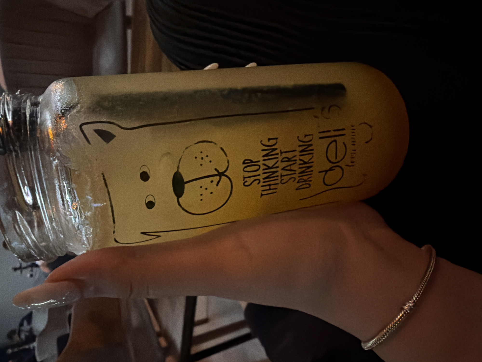 A person's hand, adorned with long, light-colored nails and a thin, gold-toned bracelet, is holding a clear glass jar filled with a yellow beverage and ice. The jar has a black drawing of a bear's face and text that reads "STOP THINKING START DRINKING deli's". The background appears to be a dimly lit indoor setting, possibly a restaurant or bar, with glimpses of seating furniture. No specific location in Plovdiv, Bulgaria, can be identified from the image.