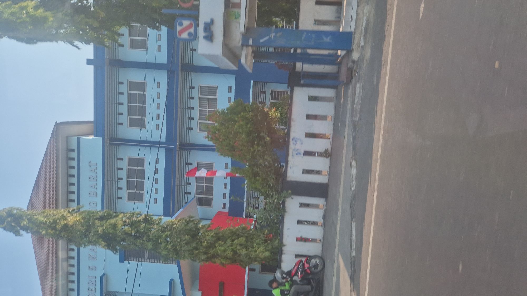 The image shows the exterior of a light blue building with a brown tiled roof, likely a school, as suggested by the text "NEGERI 5 KARAWANG BARAT" visible on the facade. The building has multiple floors with rows of windows and a blue trim. In front of the building, a white fence with rectangular openings runs along the property line. To the left, a red and white Indonesian flag is visible. A motorcycle with two individuals on it is parked in front of the fence. A tall, thin tree stands prominently in the foreground, partially obscuring the building. To the right, a structure with blue pillars and a sign that says "APL" is visible, suggesting a business or entrance to the area. The scene appears to be a street view in Karawang, Indonesia, with a clear blue sky overhead. The overall impression is a sunny day in an urban or suburban setting.