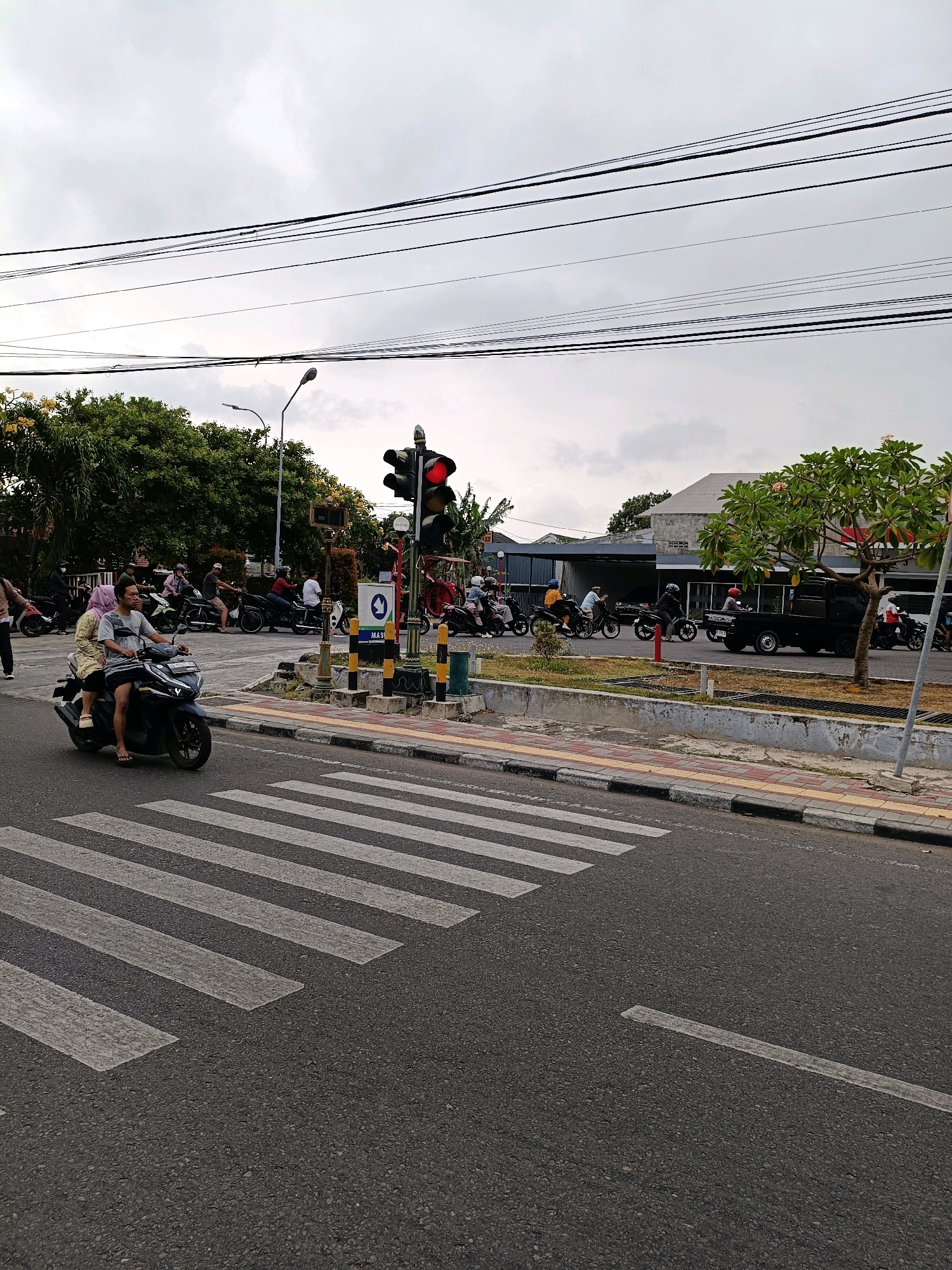 This image depicts a street scene in the city of Yogyakarta, Indonesia, likely near a road intersection. In the foreground, a pedestrian crossing is clearly visible on an asphalt road. A blue motorcycle with two occupants, a driver and a passenger wearing a headscarf, is crossing the pedestrian crossing from left to right. The driver is wearing a gray t-shirt and the passenger is wearing a patterned dress. Several other motorcycles and their riders are visible in the background, suggesting traffic is present. A traffic light at the intersection is displaying a red signal. Overhead, power lines are strung across the sky, which is overcast. Behind the traffic light, trees with dense green foliage line the side of the road. A black pick-up truck is parked or stopped on the right side of the road. Buildings, including one with a light-colored roof, are visible in the distance. The overall environment appears to be urban, with elements of road infrastructure, vehicles, and pedestrian activity.