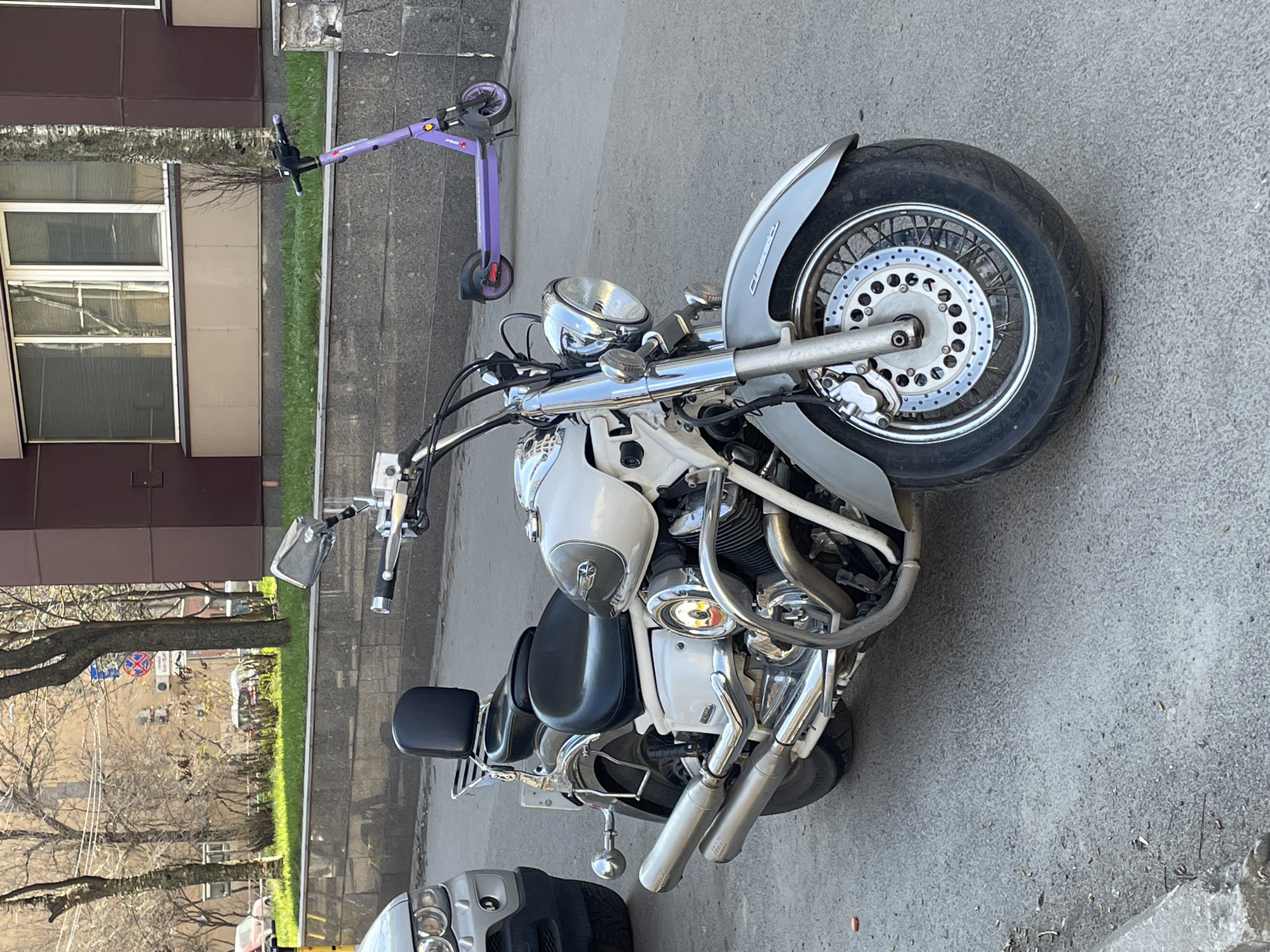 The image captures a daytime scene in Saint Petersburg, Russia, featuring a variety of parked vehicles on an asphalt surface, with urban buildings and trees in the background.
In the foreground, a prominent white and chrome cruiser motorcycle is parked. It showcases a polished chrome finish on its handlebars, headlight, engine components, exhaust pipes, and mirrors. The front fender bears the embossed word "Classic." The motorcycle's front wheel, with its spokes and disc brake system, is clearly visible. It is equipped with a black seat.
Further back, on a tiled sidewalk adjacent to a strip of green grass, a purple electric scooter is parked. The scooter's frame displays the branding "URBAN" along with an associated logo.
Partially visible in the lower left corner is the front bumper and headlights of a grey car.
The background consists of modern multi-story buildings with dark brown and beige facades, featuring numerous windows. Beyond the buildings and grass strip, a street is lined with trees that appear bare, suggesting either early spring or late autumn. A circular street sign, characterized by a red border and internal blue/white symbols, is faintly discernible through the tree branches, likely indicating traffic regulations. The overall lighting suggests a clear, bright day.