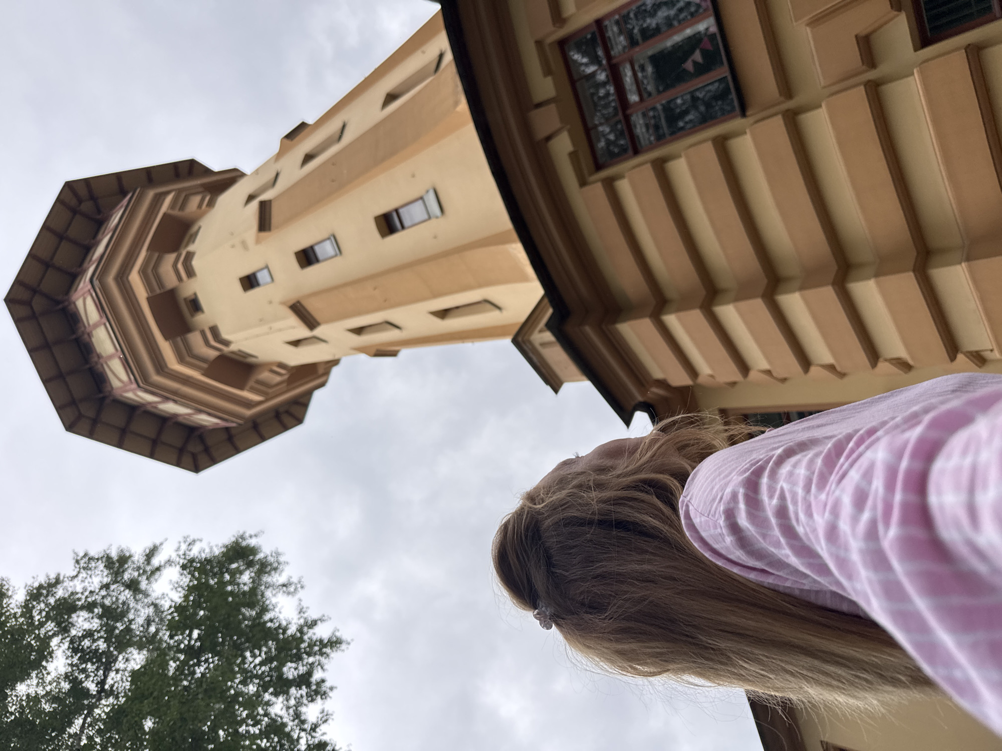 The image depicts a young woman with long blonde hair, viewed from behind and from the shoulders up, looking towards a tall, octagonal tower. She is wearing a pink and white striped shirt. The tower, painted in shades of tan and light brown, features a series of rectangular windows and decorative architectural elements. The top of the tower is crowned with a multi-tiered roof structure. The scene is set against an overcast sky, with a portion of a leafy green tree visible in the upper left corner. The building attached to the tower has a light brown facade with horizontal decorative bands and dark-framed windows. The context suggests this is the water tower located in the city of Ladozhskoe Ozero, Russia.