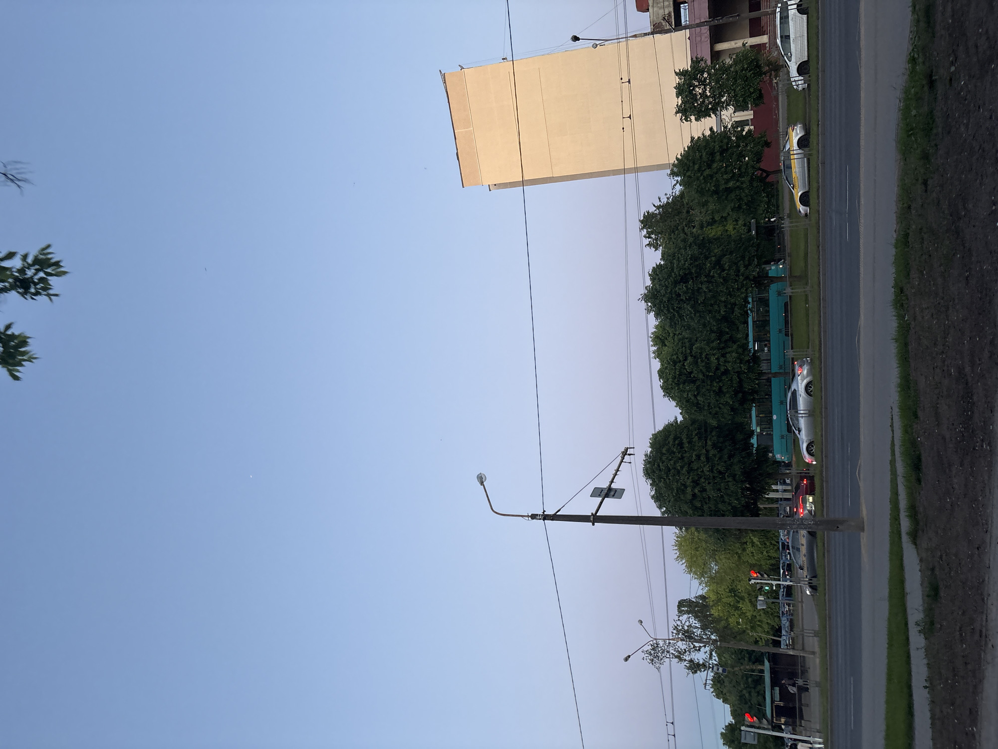 The image depicts a street scene in Minsk, Belarus. The view is from ground level, looking towards a street with a building on the left, a row of trees, a road with cars and public transport, and a sidewalk.
**Subjects and Objects:**
- Buildings: A large beige building on the left, and another building partially visible behind it.
- Trees: A line of mature trees along the sidewalk.
- Street: A paved road with multiple lanes.
- Traffic: Cars, a trolleybus, and possibly other vehicles are present in the traffic.
- Sidewalk: A paved sidewalk runs alongside the road.
- Street Furniture: Utility poles and street lights are visible.
- Sky: The sky is visible, colored in hues of blue and light purple, indicative of dusk.
- Wires: Electrical and communication wires are suspended between the poles.
**Environment and Interactions:**
- The scene is outdoors, likely in a residential or commercial area of Minsk.
- The presence of traffic indicates an urban environment.
- The time of day is likely evening or dusk, as suggested by the sky's color.
- The scene suggests ordinary urban life with transportation, infrastructure, and buildings.
**People:**
- Possible presence of people who may be waiting in public transportation at the bus station.