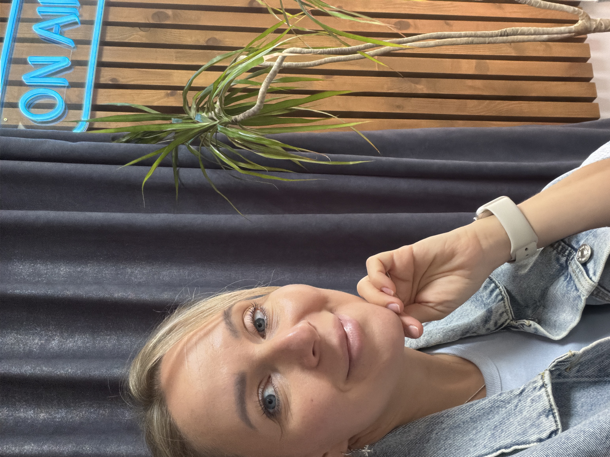 The image displays a fair-skinned woman with blonde hair and blue eyes, lying on her left side and looking directly at the camera with a soft smile. Her left hand is gently placed near her chin. She is dressed in a light blue t-shirt and a light wash denim jacket, and a light-colored smartwatch is visible on her left wrist.
The background is composed of several elements. Immediately behind the woman is a dark grey, pleated or draped fabric, likely upholstery. Above this, a slatted wooden wall creates a textured backdrop. A green, spiky-leaved plant with visible woody stems, resembling a Dracaena, extends horizontally across the frame, positioned in front of the wooden wall and above the grey fabric. In the upper left corner, a blue neon sign partially shows the illuminated letters "ON VA."
The scene appears to be an indoor setting, possibly a home or office lounge in Chaikovsky, Russia, characterized by modern decor including the wooden wall and neon signage. The lighting is soft and diffused, suggesting daytime with indirect light. The woman's relaxed posture and pleasant expression convey a sense of calm and approachability.
