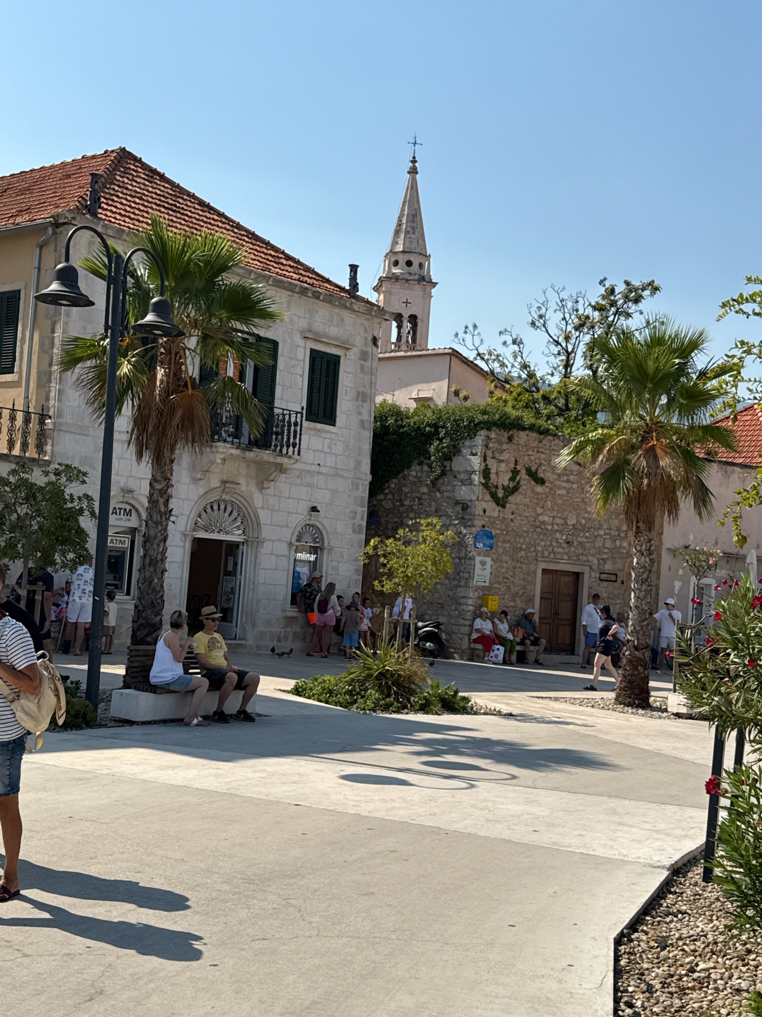 The image depicts a sunny day in Jelsa, Croatia. In the foreground, a concrete plaza is partially visible with shadows cast by palm trees and a streetlamp. A woman in a striped shirt and denim shorts is standing on the left, her back to the camera. A bench is occupied by a couple, a woman in a white tank top and shorts and a man in a yellow t-shirt and hat. Behind them, a light-colored stone building with a red-tiled roof features dark shutters and balconies. An ATM is visible on the facade of this building. Further in the background, another stone building with an arched wooden doorway stands. Palm trees are scattered throughout the scene. In the distance, a church steeple with a cross rises against a clear blue sky. People are seen walking and sitting in small groups, enjoying the pleasant weather. The overall atmosphere is one of a relaxed, pedestrian-friendly town square.